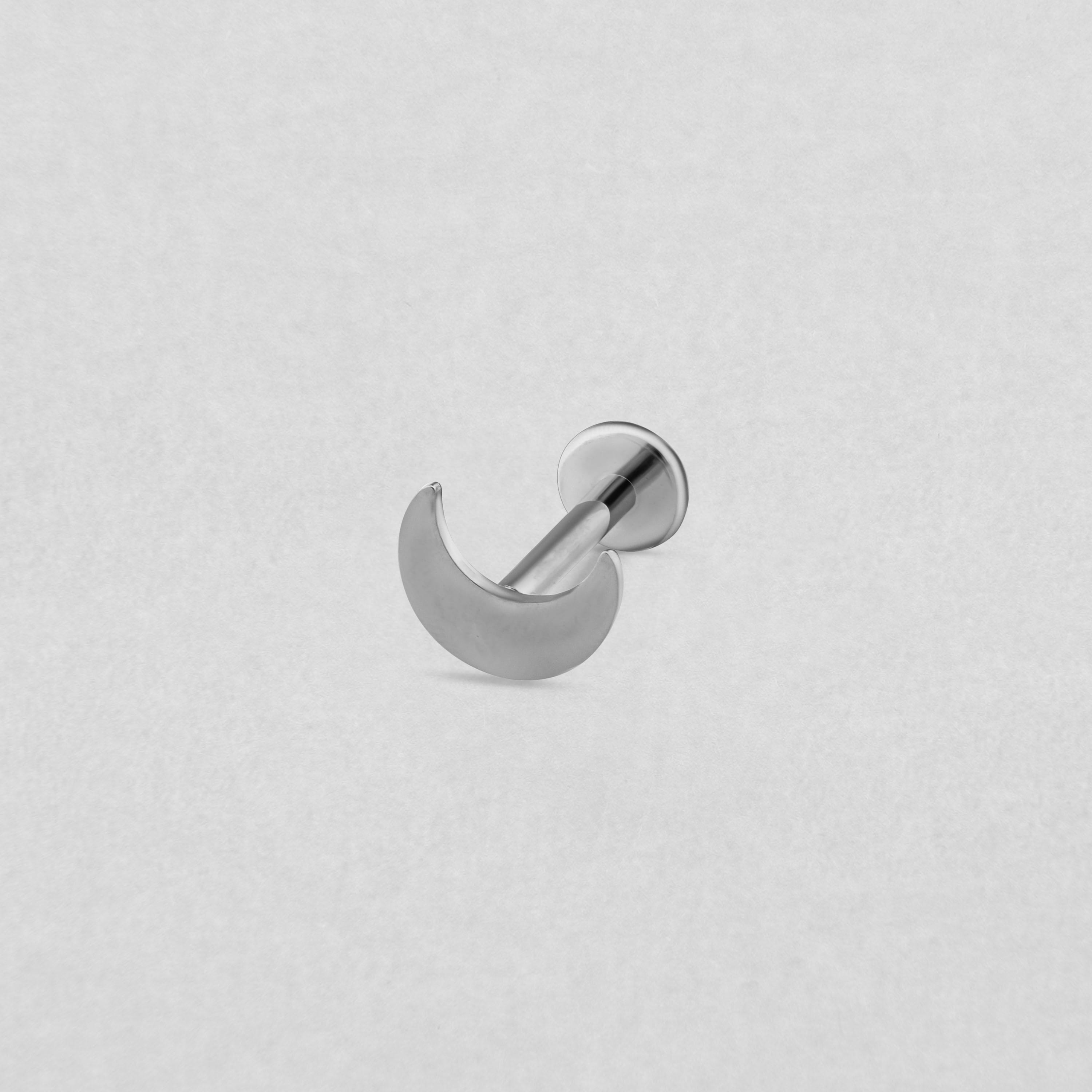 El modelo «Luna liso» de Titanio-Noble Art es un pendiente de botón con forma de media luna de plata y parte trasera plana, que destaca con elegancia sobre un fondo texturizado de color gris claro.