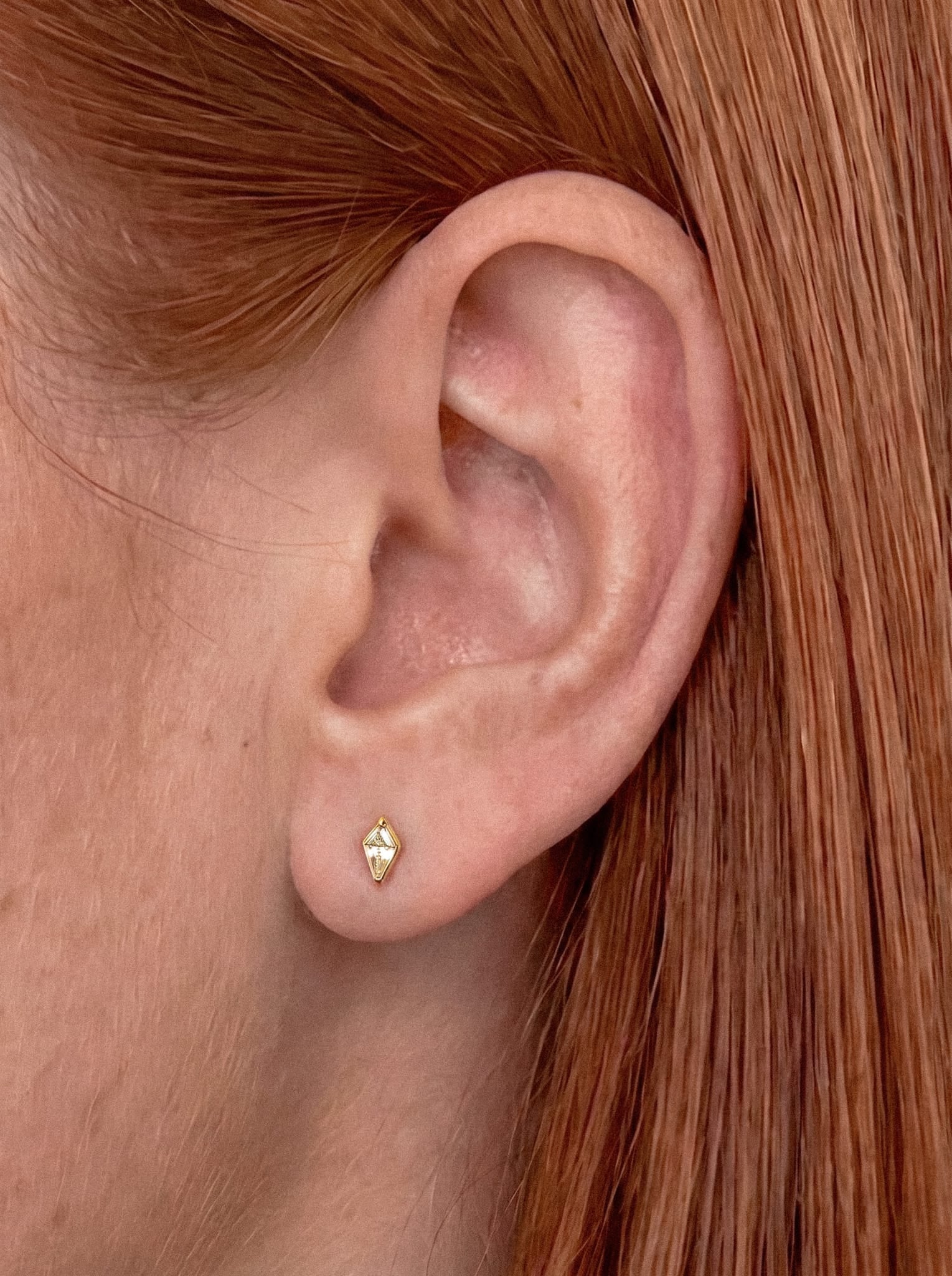 Primer plano de la oreja de una persona con el pelo liso de color castaño rojizo, que lleva el pendiente de botón geométrico dorado «Top Egan» de NOBLE ART BEAUTY SOCIEDAD LIMITADA. El pendiente presenta un estilo limpio y un diseño tipo diamante. El fondo aparece desenfocado, lo que centra la atención en la oreja y en el elegante pendiente.