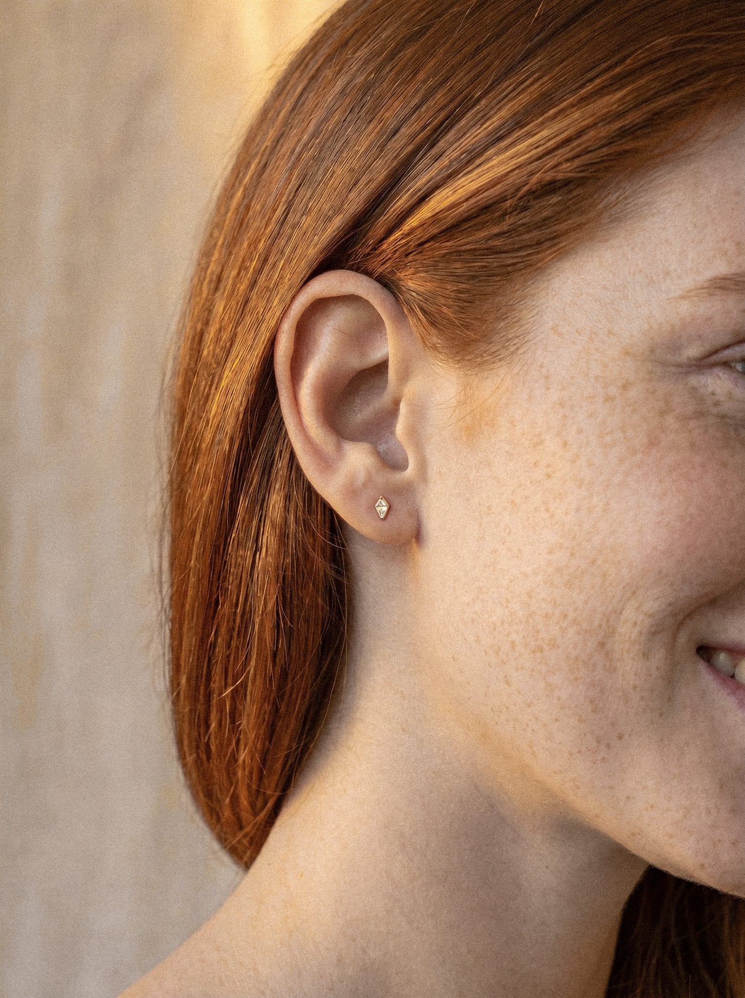 Un primer plano de una persona sonriente con el pelo pelirrojo, en el que se aprecia el pendiente de botón «Top Egan» de NOBLE ART BEAUTY SOCIEDAD LIMITADA, con un diseño inspirado en los diamantes que le confiere un aspecto sencillo y elegante.