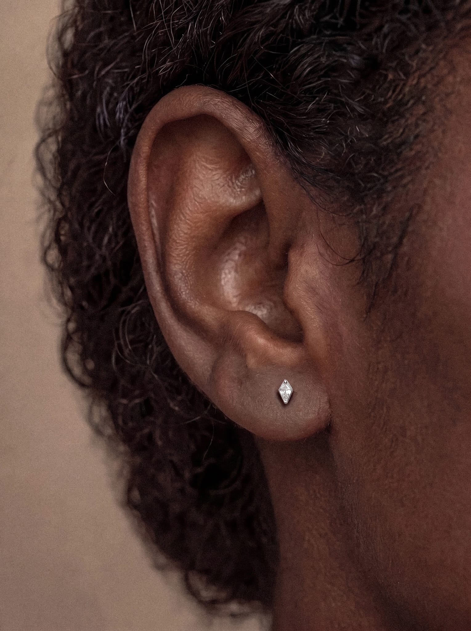 Primer plano de la oreja de una persona con el pelo corto y rizado, que lleva en el lóbulo el pendiente de botón «Top Egan» de NOBLE ART BEAUTY SOCIEDAD LIMITADA. El diseño en forma de diamante aporta un toque de estilo limpio al look, y esta pieza está fabricada en acero resistente para garantizar una elegancia duradera.