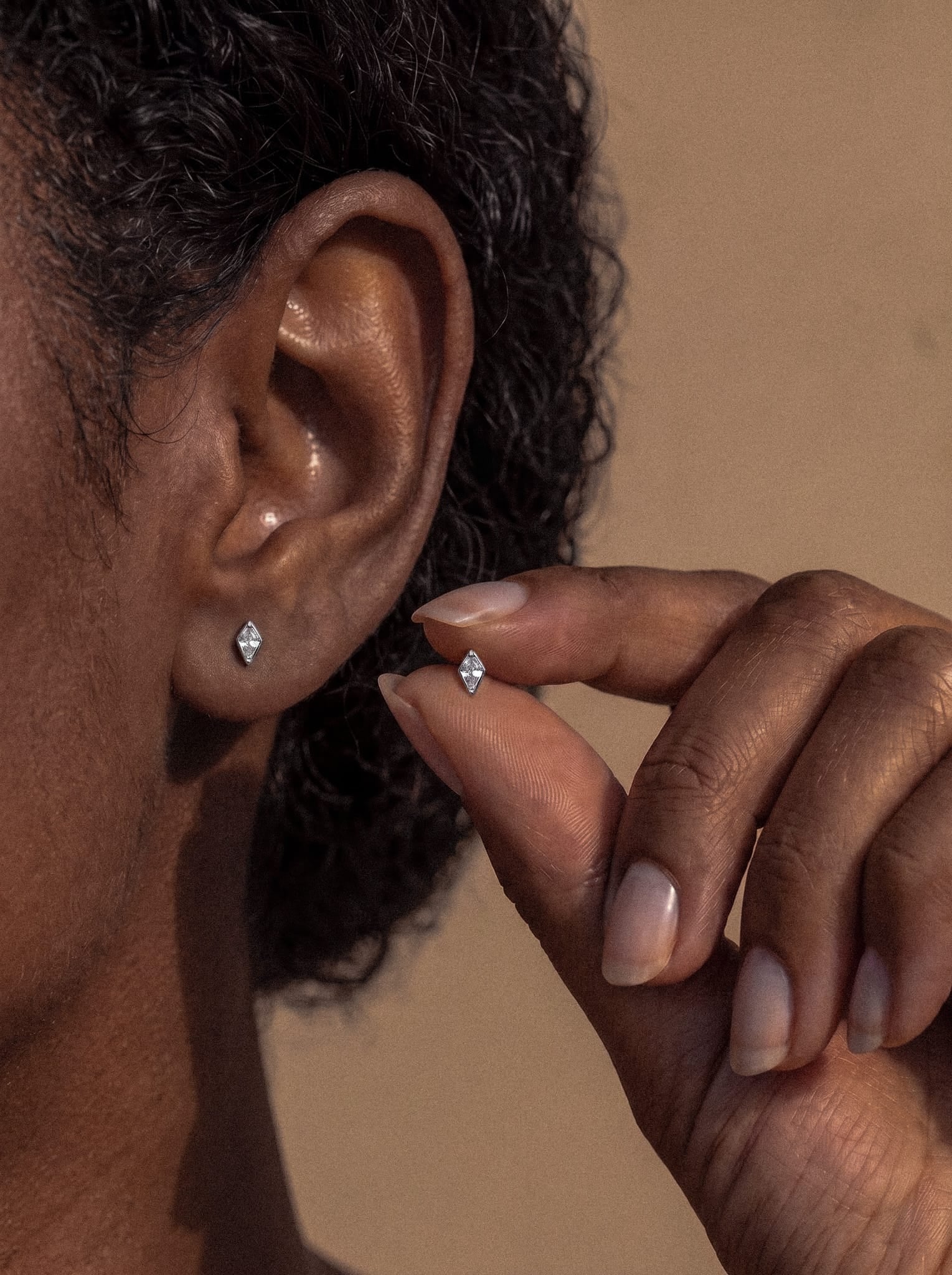 Primer plano de la oreja de una persona que lleva el pendiente tipo diamante «Top Egan» de NOBLE ART BEAUTY SOCIEDAD LIMITADA, con la mano sosteniendo otro pendiente similar junto a la oreja, sobre un fondo neutro que resalta su diseño limpio y elegante.