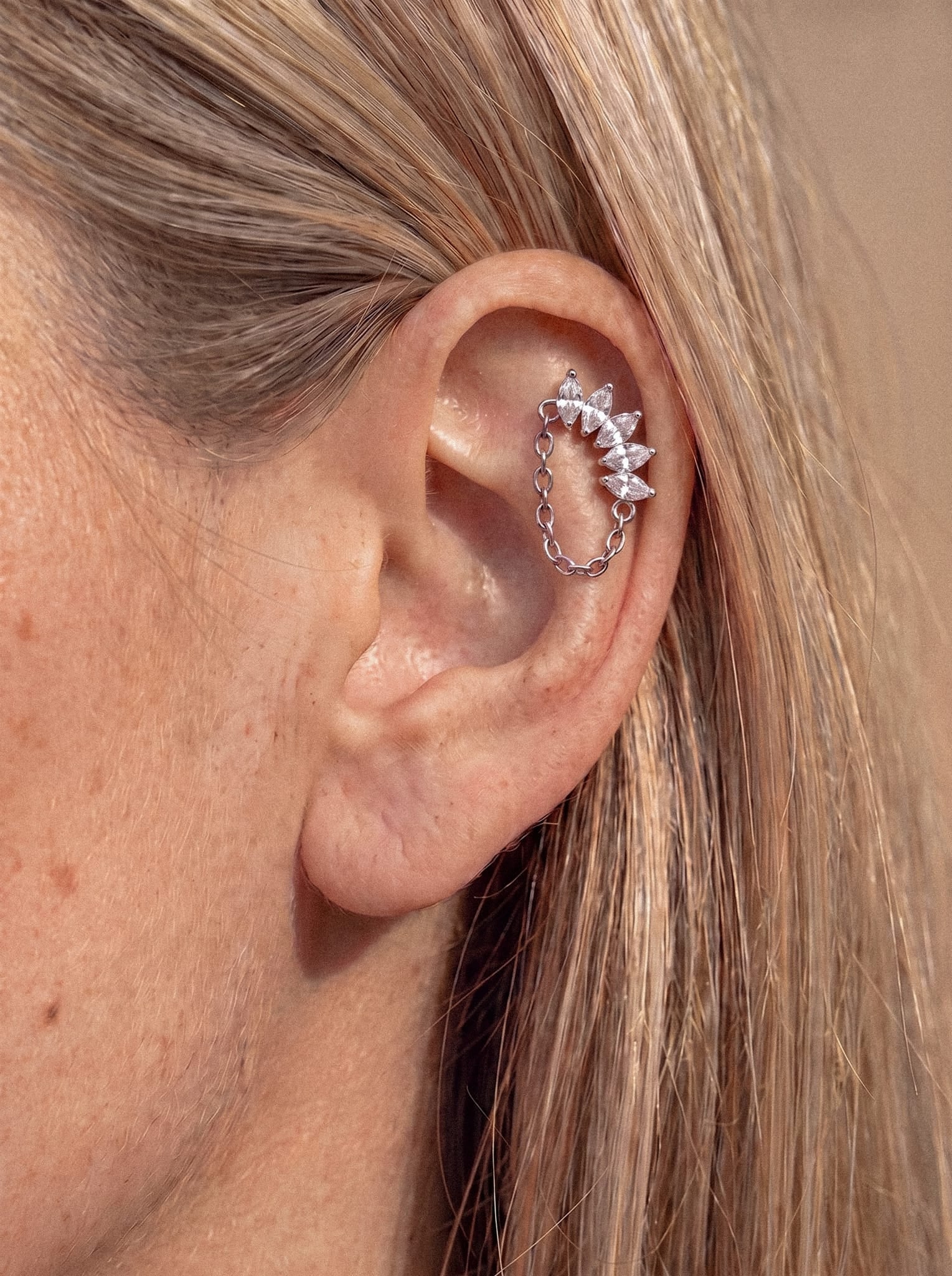 Primer plano de la oreja de una persona en el que se aprecia un peinado de moda con el pendiente tipo «ear cuff» Electra de plata de Titanio-Noble Art, adornado con varias gemas pequeñas y puntiagudas de talla marquesa y una delicada cadena, lo que aporta un sutil toque de inspiración rockera sobre un fondo neutro. La persona tiene el pelo rubio claro.