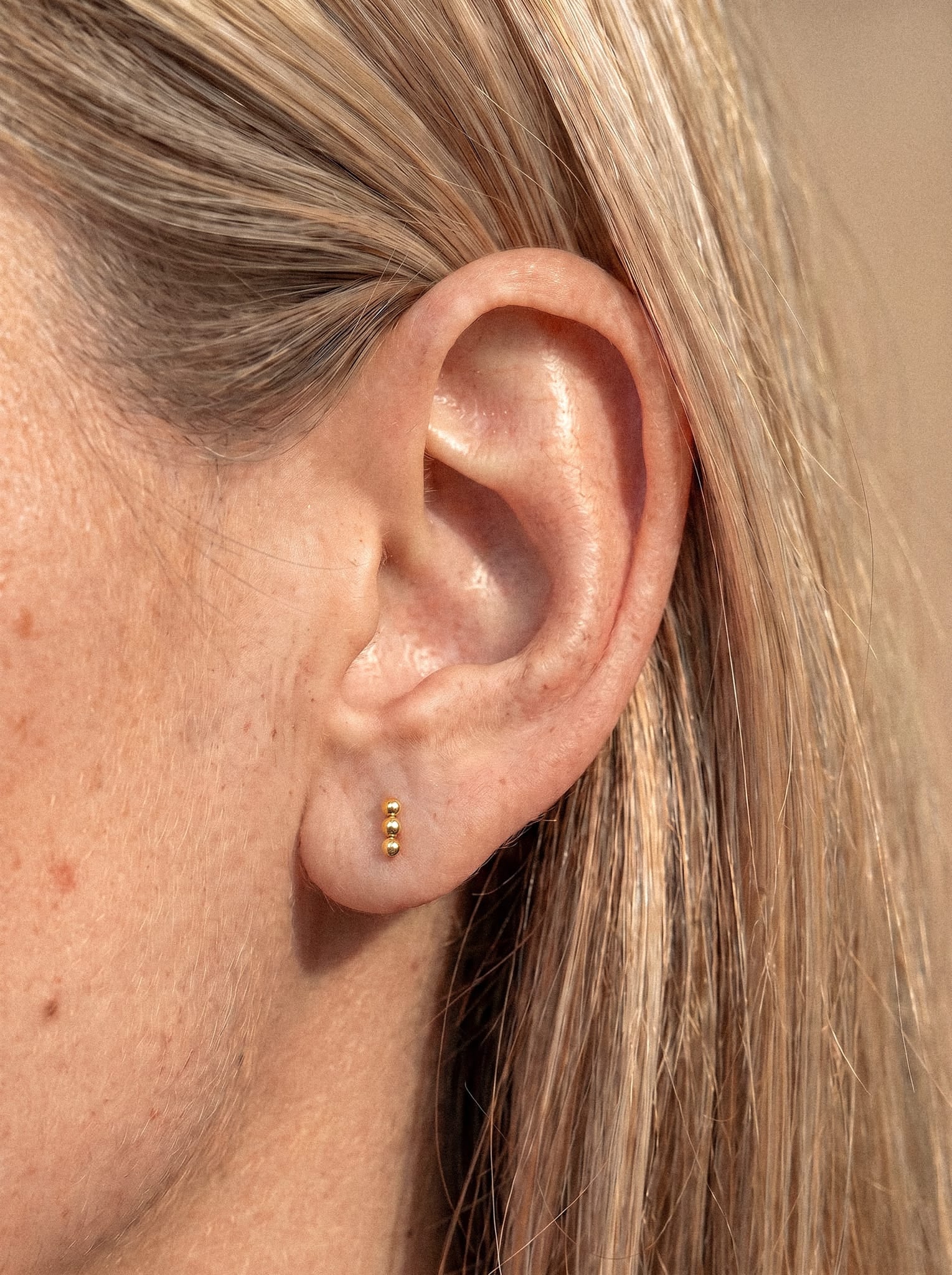 Primer plano de la oreja de una persona con tres pequeños pendientes de botón plateados «Fenix» de Titanio-Noble Art, alineados verticalmente en el lóbulo para crear un efecto de simetría metálica; la persona tiene la piel clara y el pelo rubio, lo que resalta el diseño minimalista de este elegante look.