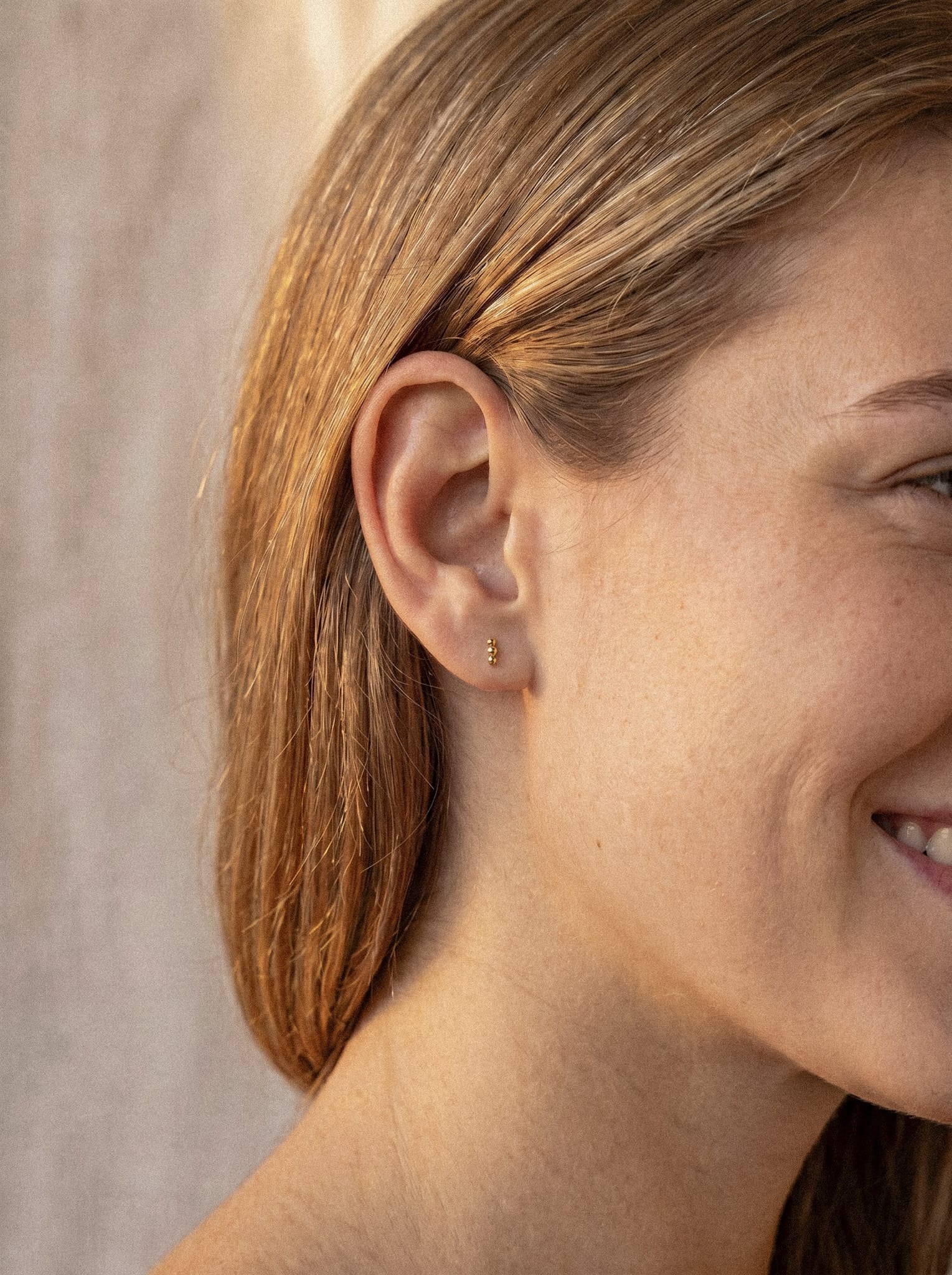 Primer plano de la oreja de una mujer sonriente, con el pelo liso de color castaño claro, que lleva el pendiente de aro plateado «Fenix» de Titanio-Noble Art, que destaca por su diseño sutil y minimalista. El fondo es neutro y está suavemente iluminado.