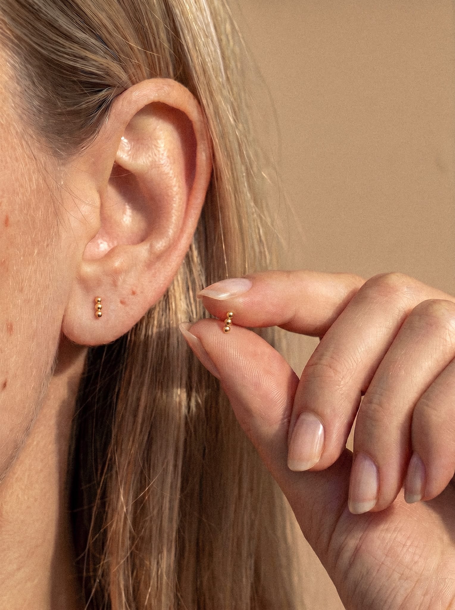 Un primer plano de la oreja de una persona con un pequeño pendiente de botón de plata «Fenix» de Titanio-Noble Art, que destaca por su sutil diseño minimalista. La persona sujeta entre el pulgar y el índice, cerca de la oreja, un pendiente de botón de plata «Fenix» a juego, lo que resalta la simetría metálica de los accesorios.