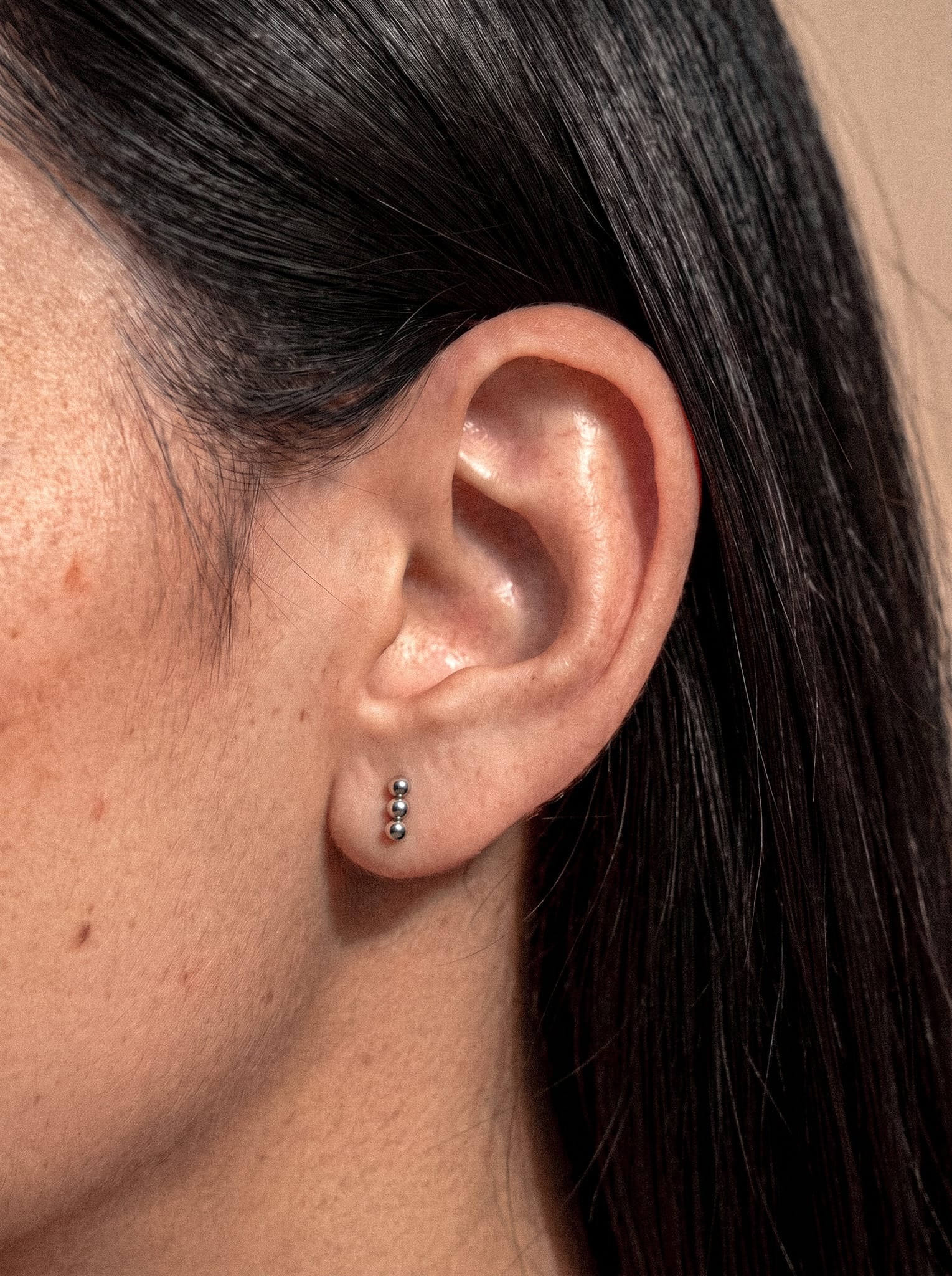 Un primer plano de la oreja de una persona con el pelo liso y oscuro, adornada con el pendiente de botón de plata «Fénix» de Titanio-Noble Art, que presenta tres bolas de plata dispuestas verticalmente en el lóbulo de la oreja, creando un efecto de simetría metálica y un diseño minimalista y evocador.