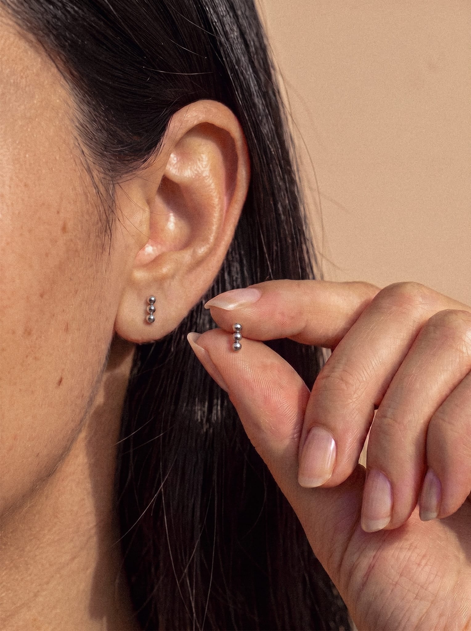 Un primer plano de la oreja de una persona, adornada con tres pequeños pendientes de bola de plata «Fenix» de Titanio-Noble Art, muestra un diseño minimalista impecable y una simetría metálica; la persona sujeta un pendiente a juego entre el pulgar y el índice.