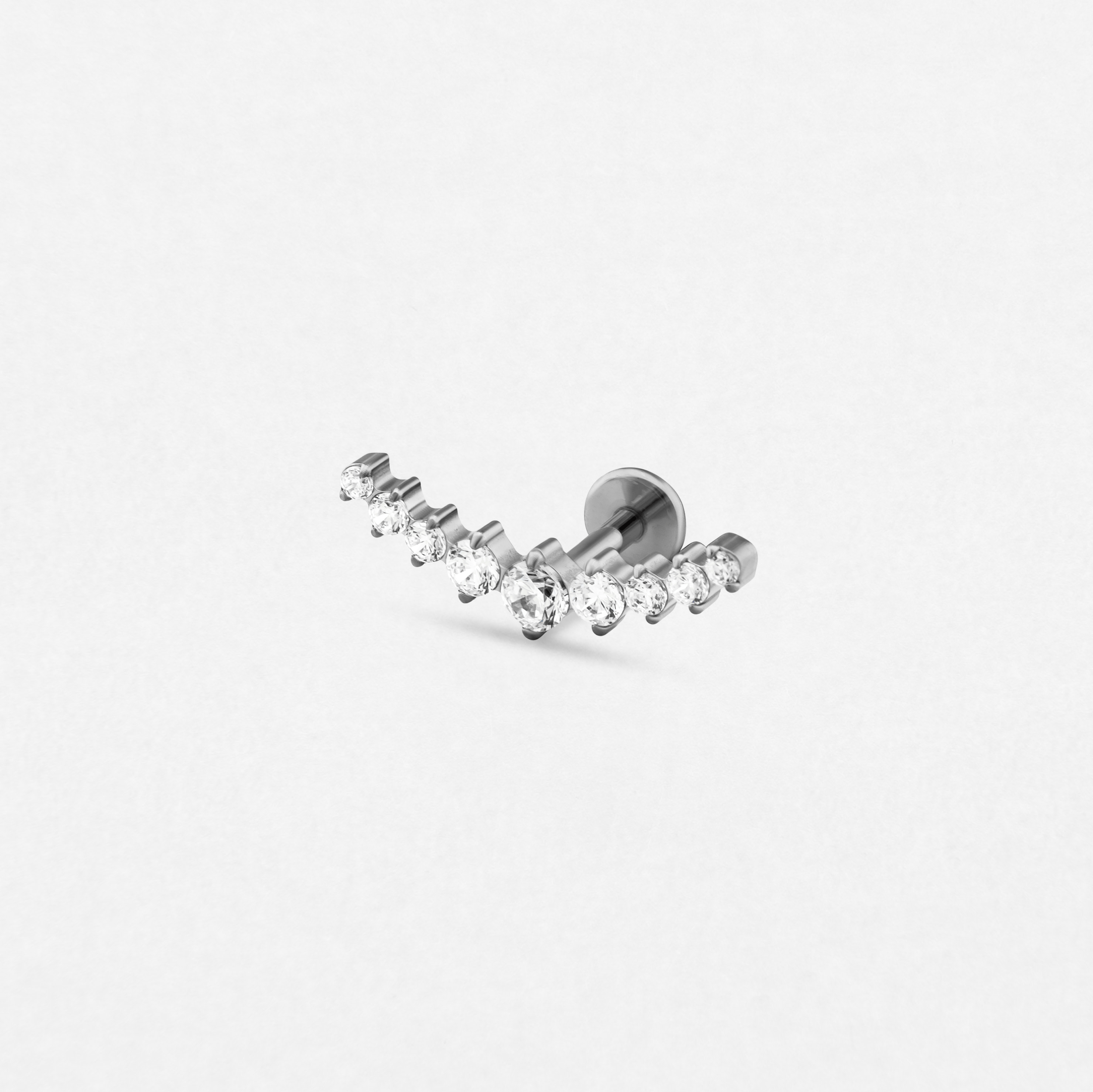 El pendiente tipo «ear cuff» Leo de Titanio-Noble Art presenta un diseño sencillo en plata con pequeñas piedras preciosas redondas y transparentes dispuestas en una suave curva, sobre un fondo blanco liso.