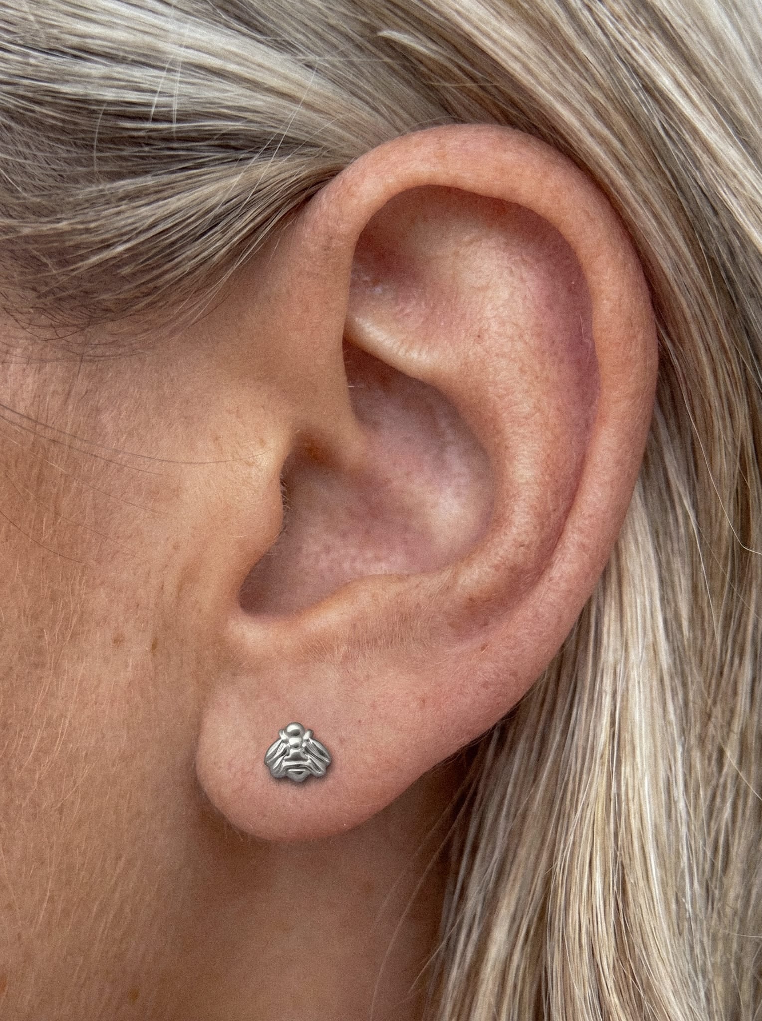 Eine Nahaufnahme des Ohrs einer Person mit dem Ohrring „Lisa“ von Titanio-Noble Art – einem kleinen, gehirnförmigen Silberstecker –, der sich vor heller Haut und blonden Haaren abhebt: das perfekte minimalistische Piercing für alle, die auf der Suche nach einem originellen Geschenk sind.