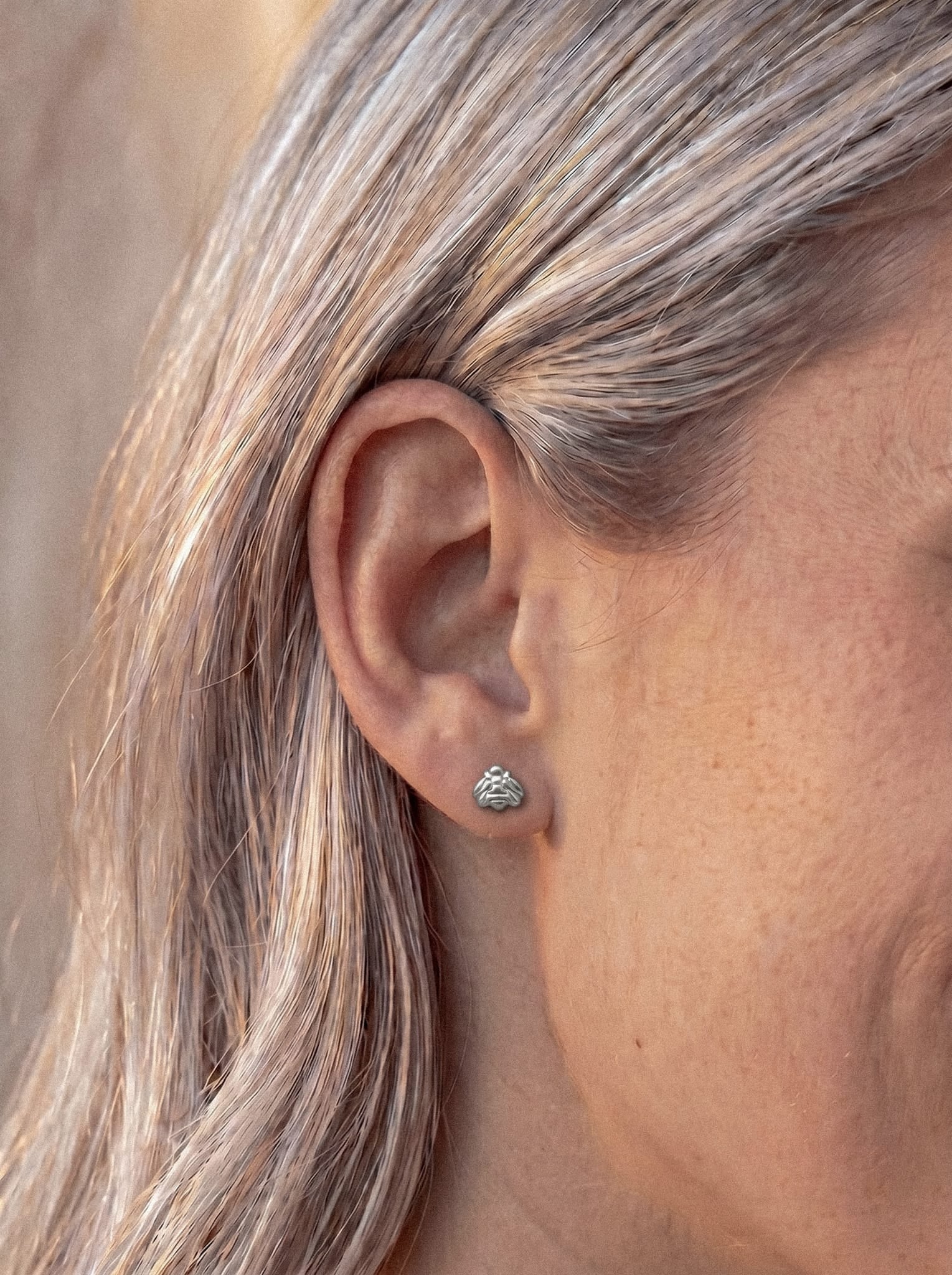 Nahaufnahme des Ohrs einer Person mit glattem, hellem Haar, die „Lisa“ trägt – einen kleinen Metall-Ohrstecker von Titanio-Noble Art in Form eines eleganten Wellenmusters – ein ideales Beispiel für „Piercing Minimalista“. Der sanft unscharfe Hintergrund unterstreicht die raffinierte Schlichtheit dieses goldenen Schmuckstücks und macht es zu einem perfekten, originellen Geschenk.