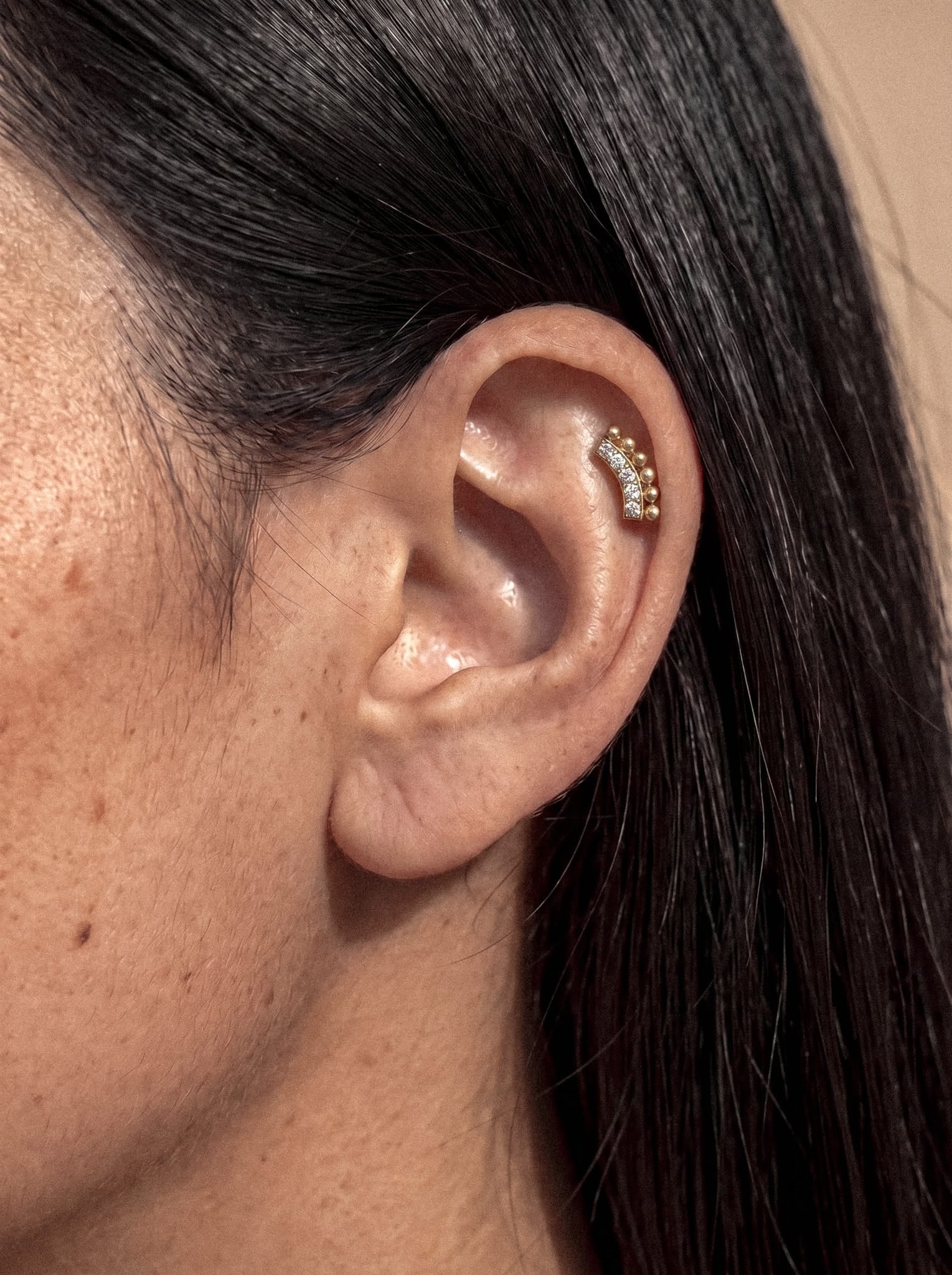 Primer plano de la oreja de una persona adornada con el pendiente «Luli» de Titanio-Noble Art, que presenta un diseño curvo de oro con esferas doradas y pequeños cristales a lo largo del cartílago interno. La persona tiene el pelo liso y oscuro y la piel suave.
