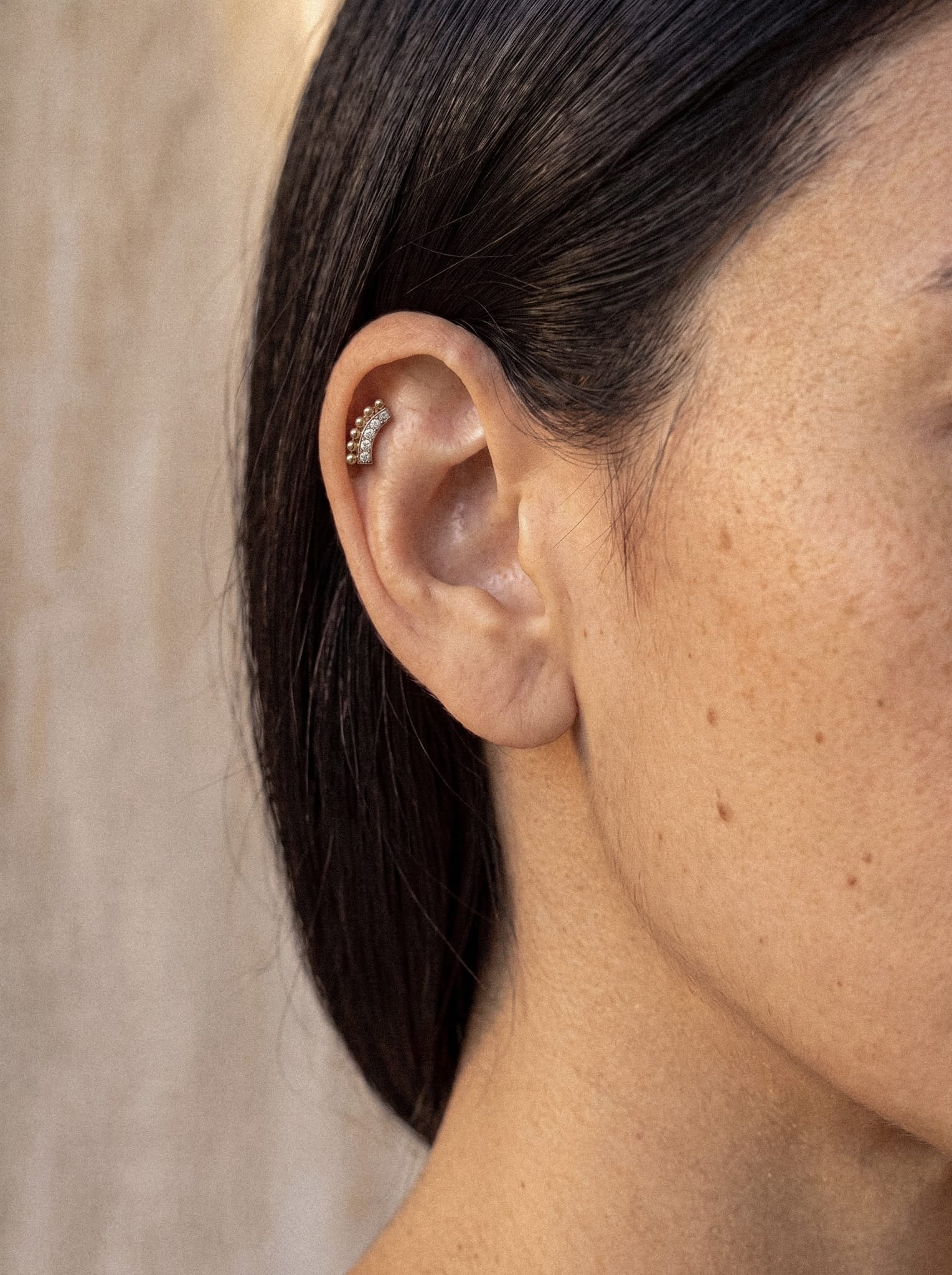 Eine Nahaufnahme zeigt das Ohr einer Frau, geschmückt mit dem „Ear Cuff“-Ohrring „Luli“ von Titanio-Noble Art, dessen zarte Kristalle auf ihrem Ohrknorpel funkeln. Ihr dunkles Haar ist sorgfältig hinter dem Ohr zusammengebunden, wodurch ihre zarte, mit einigen Sommersprossen übersäte Haut vor einem neutralen, leicht unscharfen Hintergrund besonders gut zur Geltung kommt.