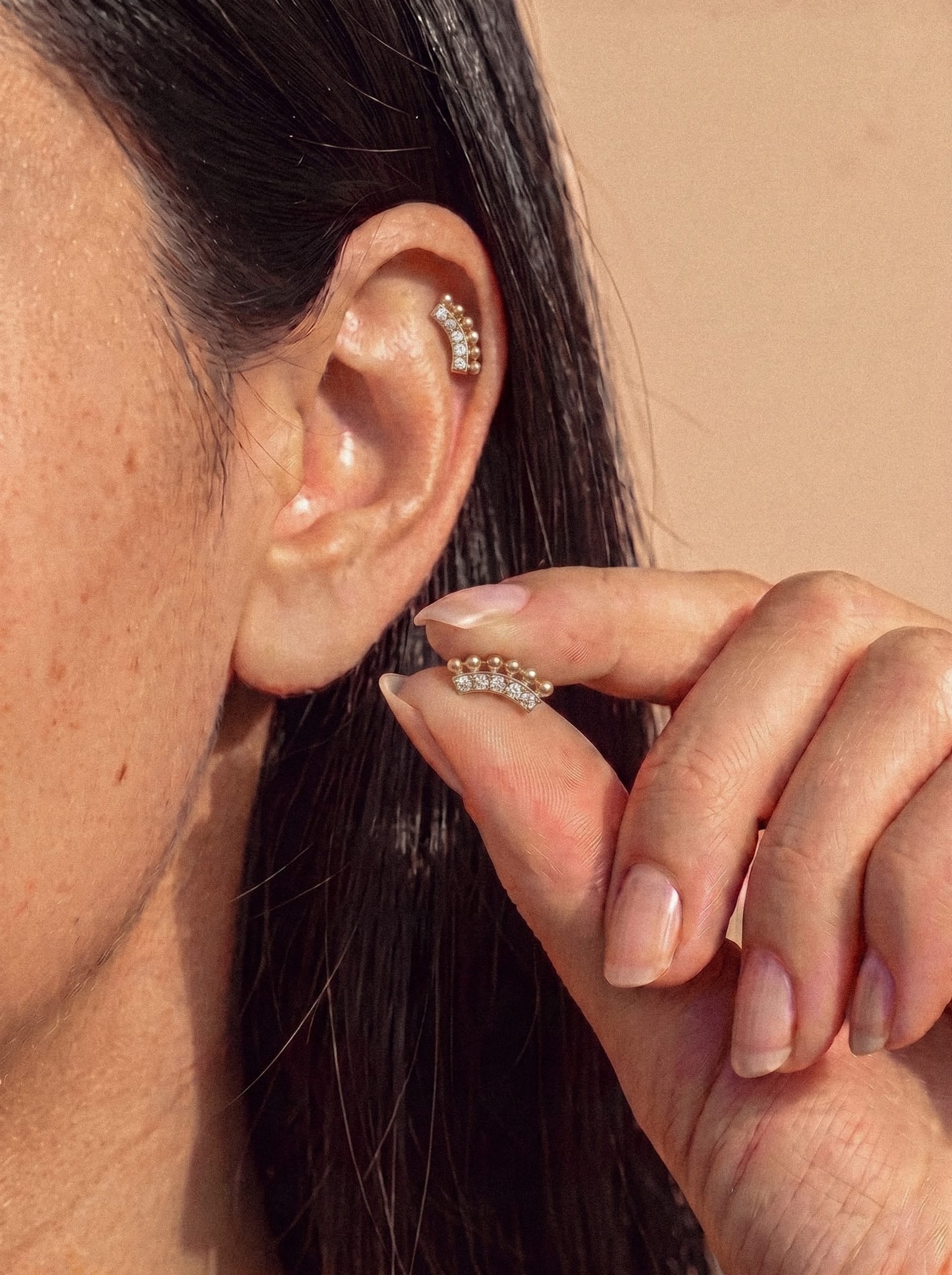 Eine Person hält den Ohrring vom Typ „Ear Cuff“ Luli aus Titan – Noble Art – an ihr Ohr – ein goldenes, geschwungenes Accessoire, das mit Perlen und goldenen Kugeln verziert ist –, während sie am Ohr bereits einen passenden Ohrring trägt. Der Hintergrund ist in einem neutralen Beigeton gehalten.