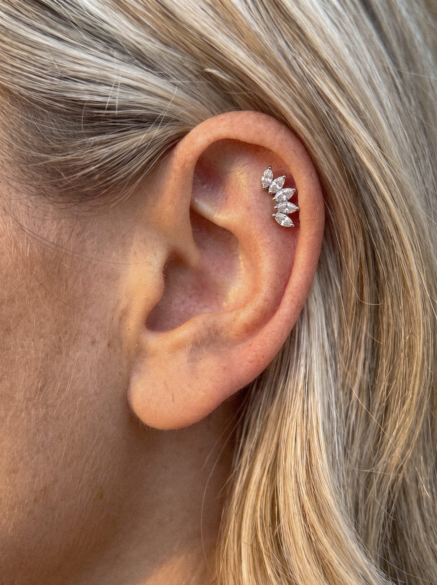 Gros plan sur l'oreille d'une personne aux cheveux blonds, qui porte la boucle d'oreille de type « ear cuff » en argent « Titanio-Noble Art Marquise 5 cz », ornée de cinq pierres précieuses taillées en « marquise » en forme de feuille le long du bord extérieur : un accessoire élégant qui s'harmonise à la perfection avec les looks modernes.