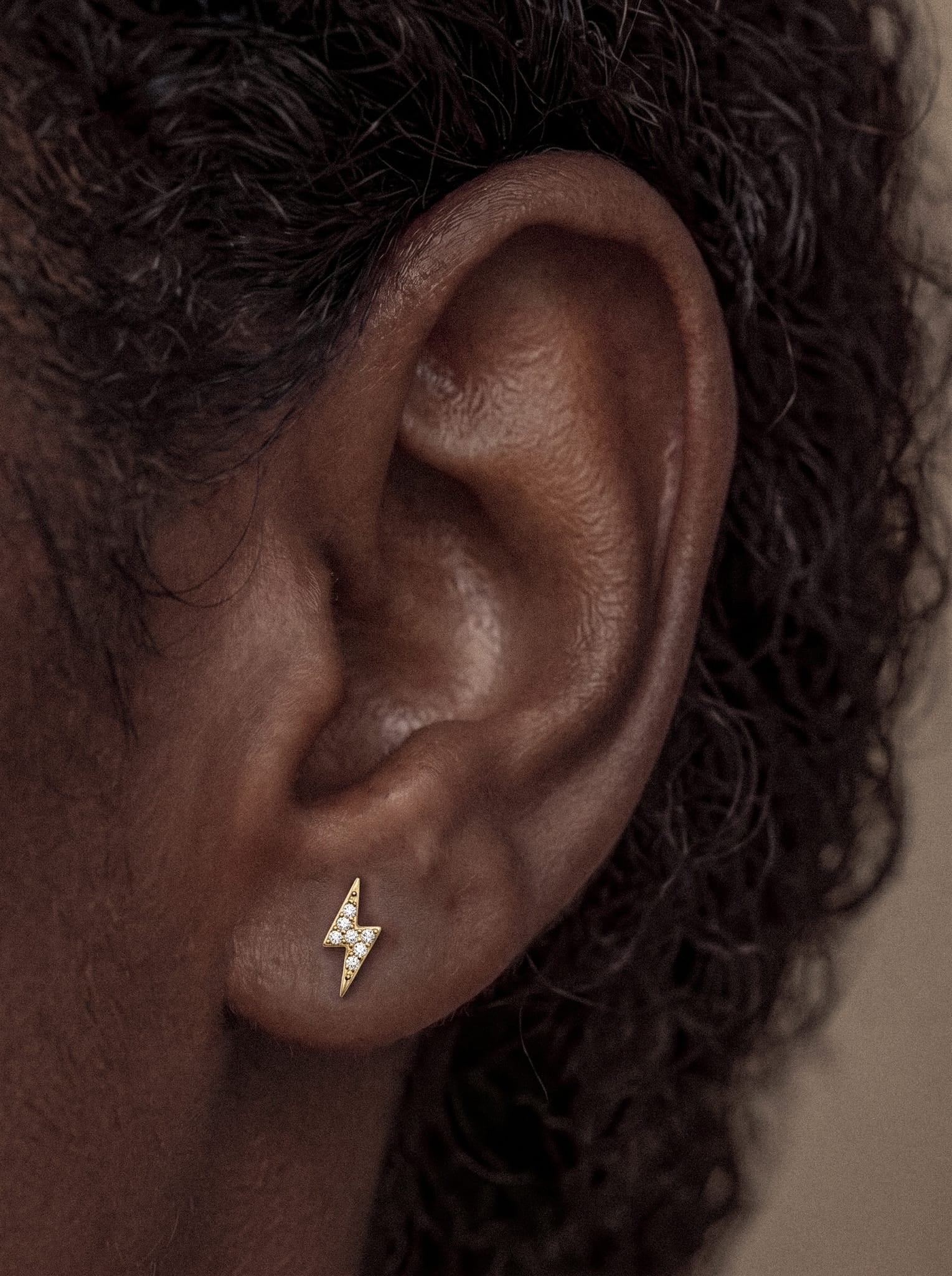 Primer plano de la oreja de una persona adornada con el pendiente de oro Maya con forma de rayo de la marca Titanio-Noble Art, que recuerda a un rayo. La persona tiene el pelo oscuro y rizado, sobre un fondo neutro.