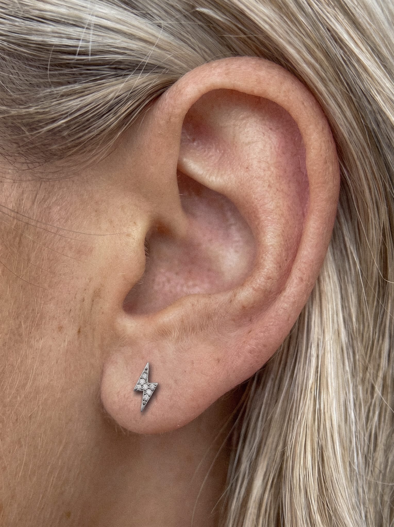 Un primer plano de una oreja con el pelo rubio y liso muestra a Maya, un pequeño pendiente plateado con forma de rayo de la marca Titanio-Noble Art, delicadamente adornado con circonitas cúbicas: un complemento perfecto para un piercing que aporta un toque de brillo sutil.