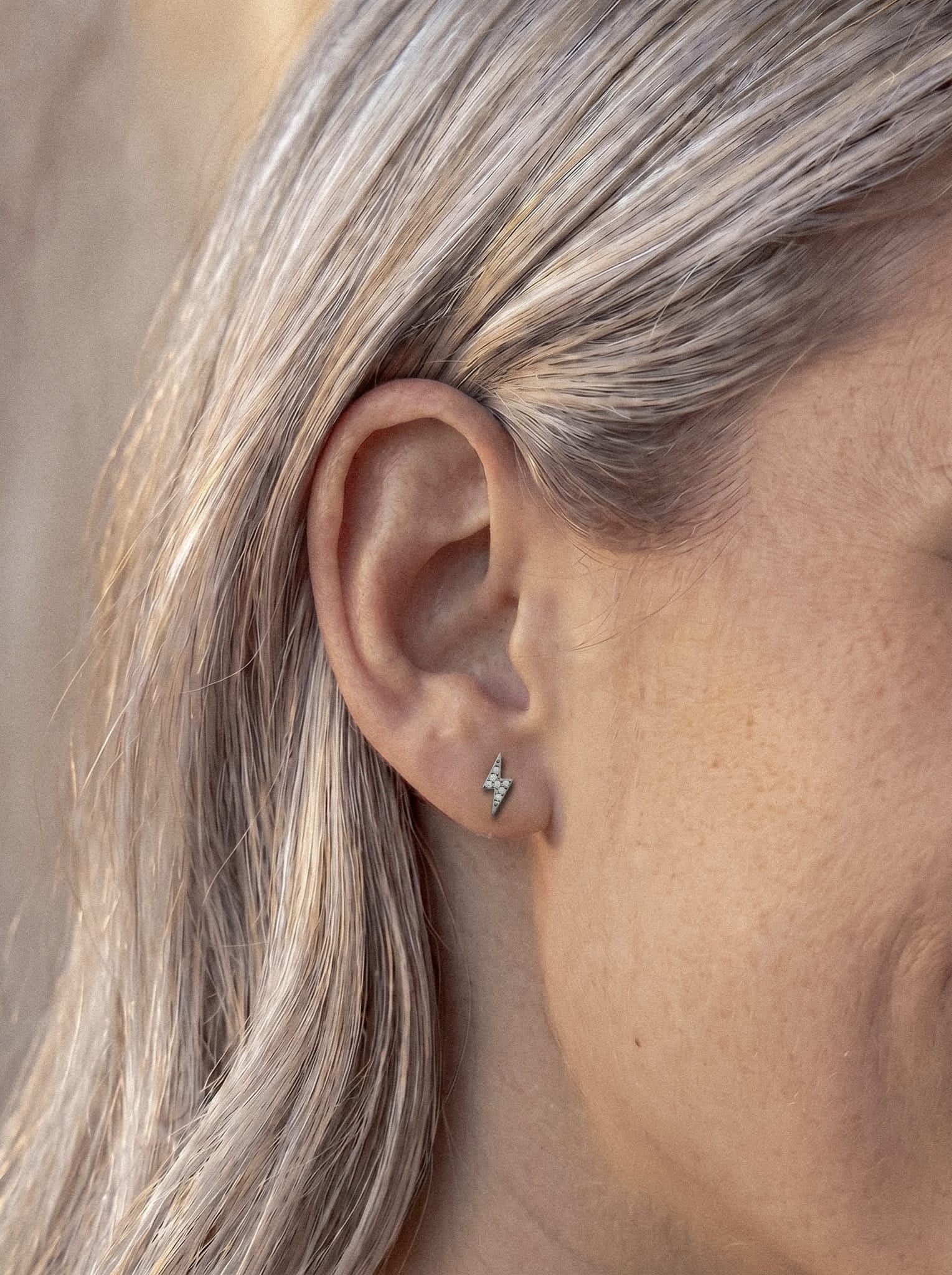 Un primer plano de la oreja de una persona con el pelo largo y rubio claro muestra a Maya, un pequeño pendiente plateado con forma de rayo de la marca Titanio-Noble Art, delicadamente engastado con circonitas cúbicas para darle un brillo extra.