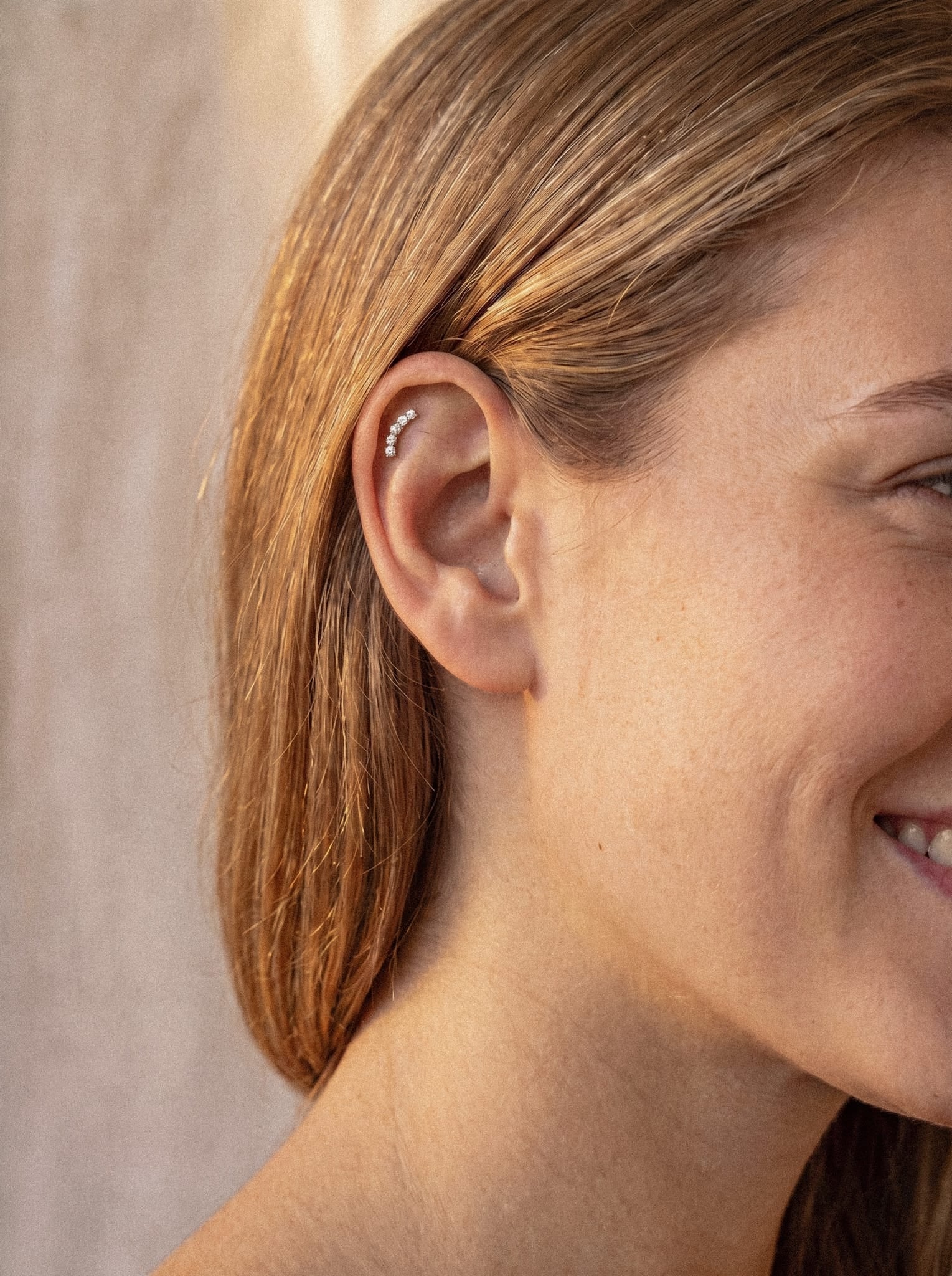 Primer plano del rostro sonriente de una mujer, con el foco puesto en su oreja, adornada con el pendiente «Mia» de Titanio-Noble Art: un diseño delicado y curvo con pequeños cristales vibrantes. Su pelo liso y castaño claro está recogido detrás de la oreja, lo que resalta los elegantes tonos joya de esta pieza de joyería.
