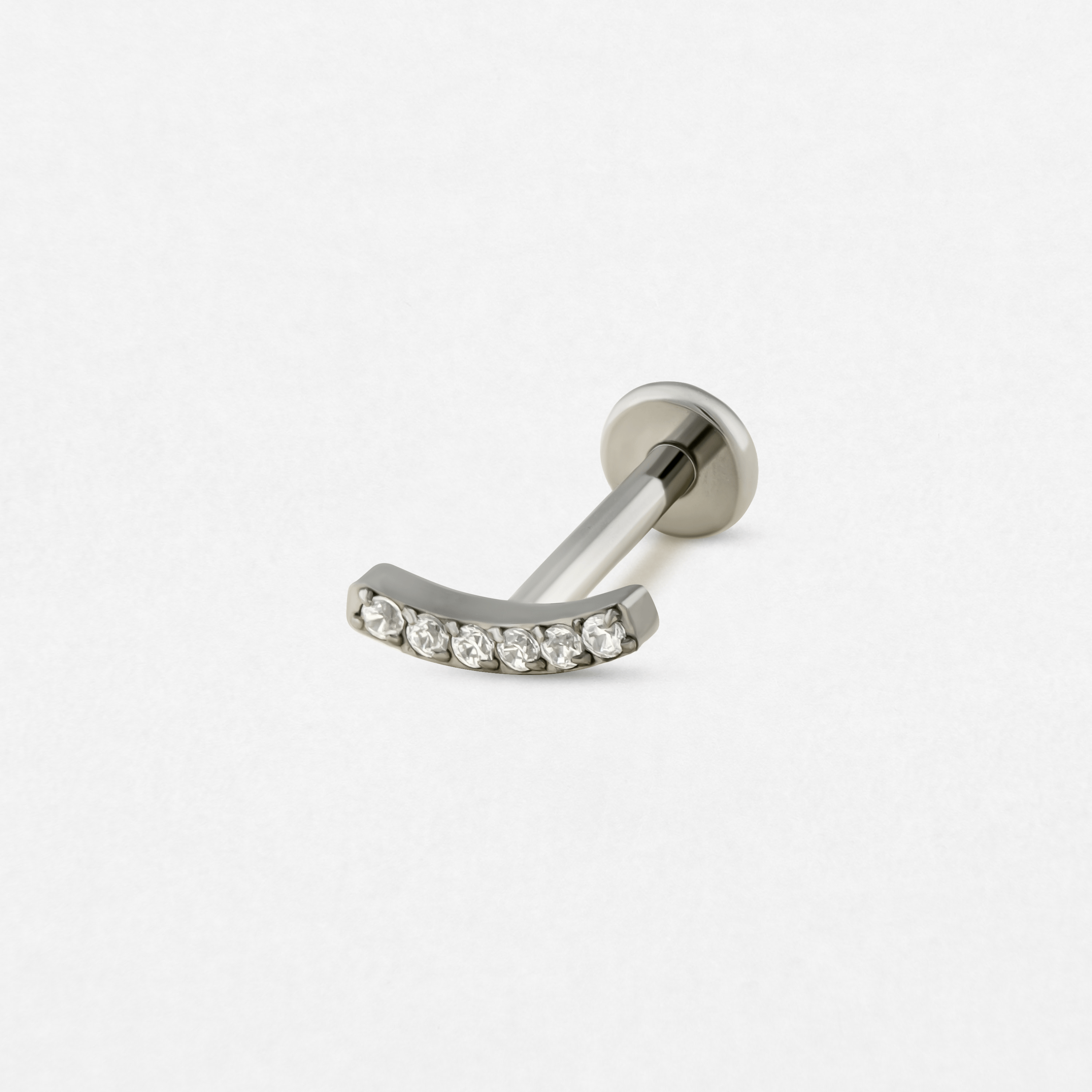 La barra curvada de plata «Mia» de Titanio-Noble Art cuenta con siete piedras preciosas transparentes engastadas a lo largo de su arco, que destacan sobre un fondo blanco liso.
