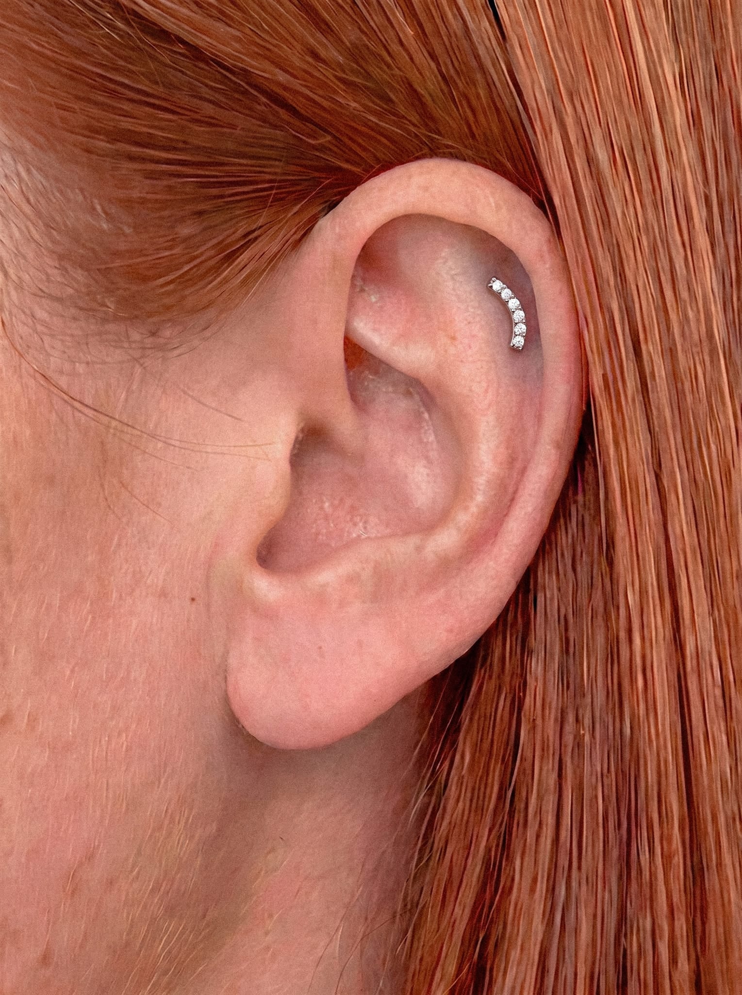 Un primer plano de la oreja de una persona adornada con los pendientes «Mia» de Titanio-Noble Art —una fila curvada de pequeños pendientes de botón brillantes en el cartílago— que muestran un piercing «delicado». La persona tiene el pelo liso, de color castaño rojizo.