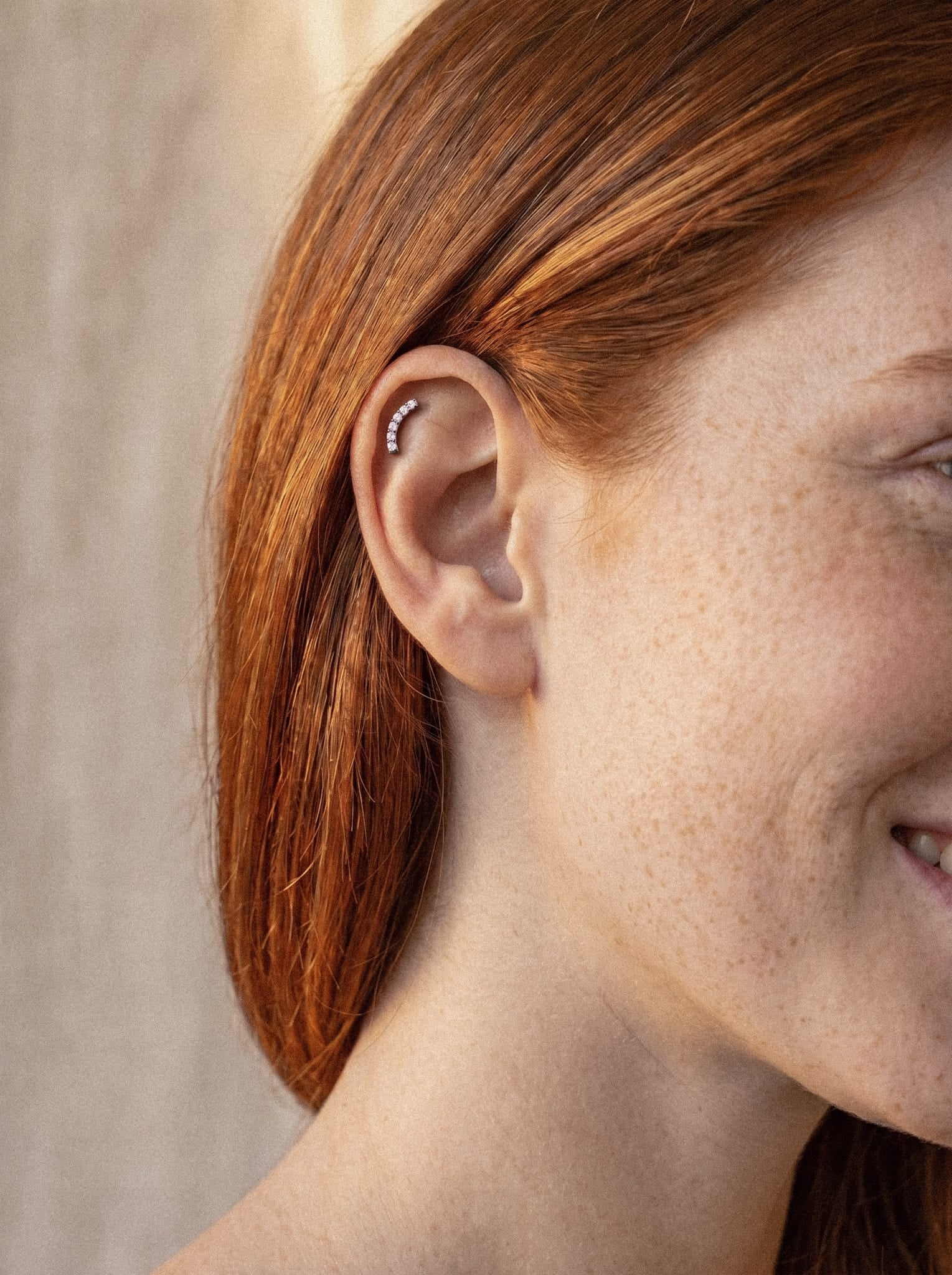 Un primer plano de una persona sonriente con el pelo largo y pelirrojo, luciendo el modelo «Mia» de Titanio-Noble Art: un pendiente de oreja curvo de plata adornado con pequeñas piedras preciosas en la parte superior de la oreja, un elegante ejemplo de piercing delicado y femenino.