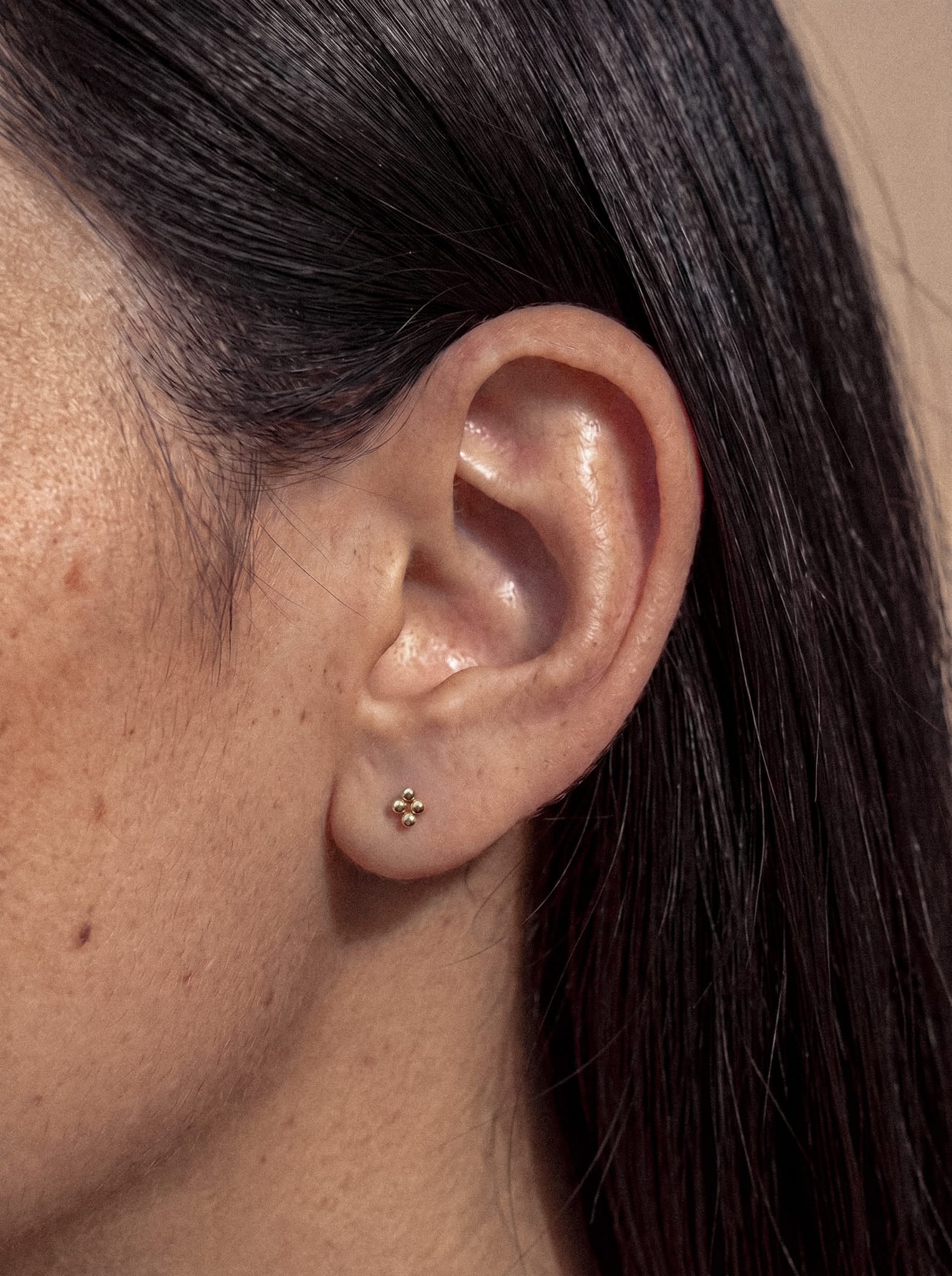 Primer plano de la oreja de una persona con el pelo liso y oscuro, que lleva el pendiente pequeño de oro «Top Nido», con forma de cruz, de NOBLE ART BEAUTY SOCIEDAD LIMITADA. Este delicado accesorio aporta un toque elegante a su estilo, en contraste con un fondo sencillo y neutro.