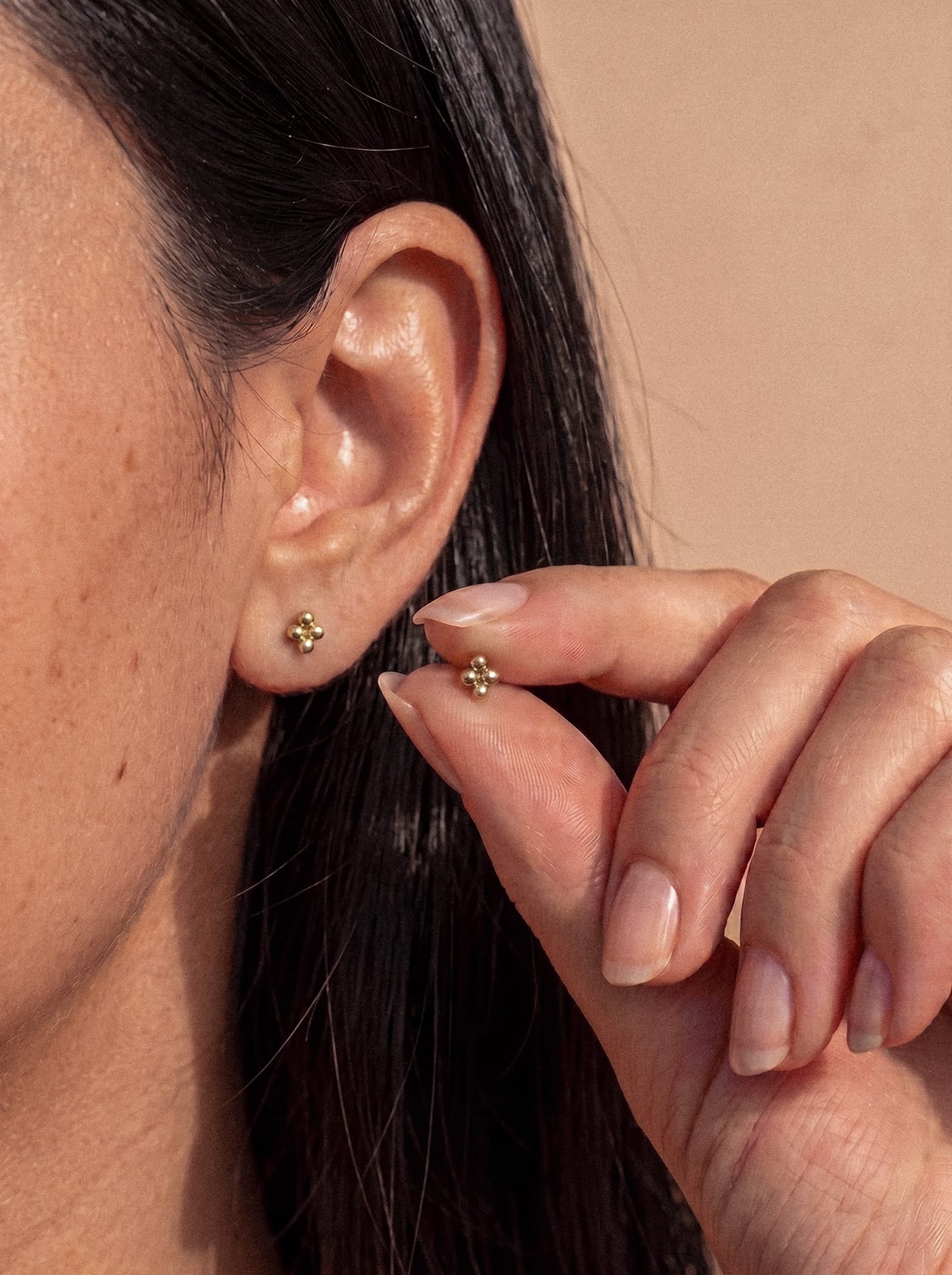 Un primer plano de la oreja de una mujer que luce el pendiente «Top Nido» de NOBLE ART BEAUTY SOCIEDAD LIMITADA: un pequeño pendiente de oro con forma de tres esferas doradas unidas entre sí. Sostiene entre los dedos, cerca de la oreja, una pieza a juego, lo que resalta su aspecto delicado y el estilo de los pendientes modernos.