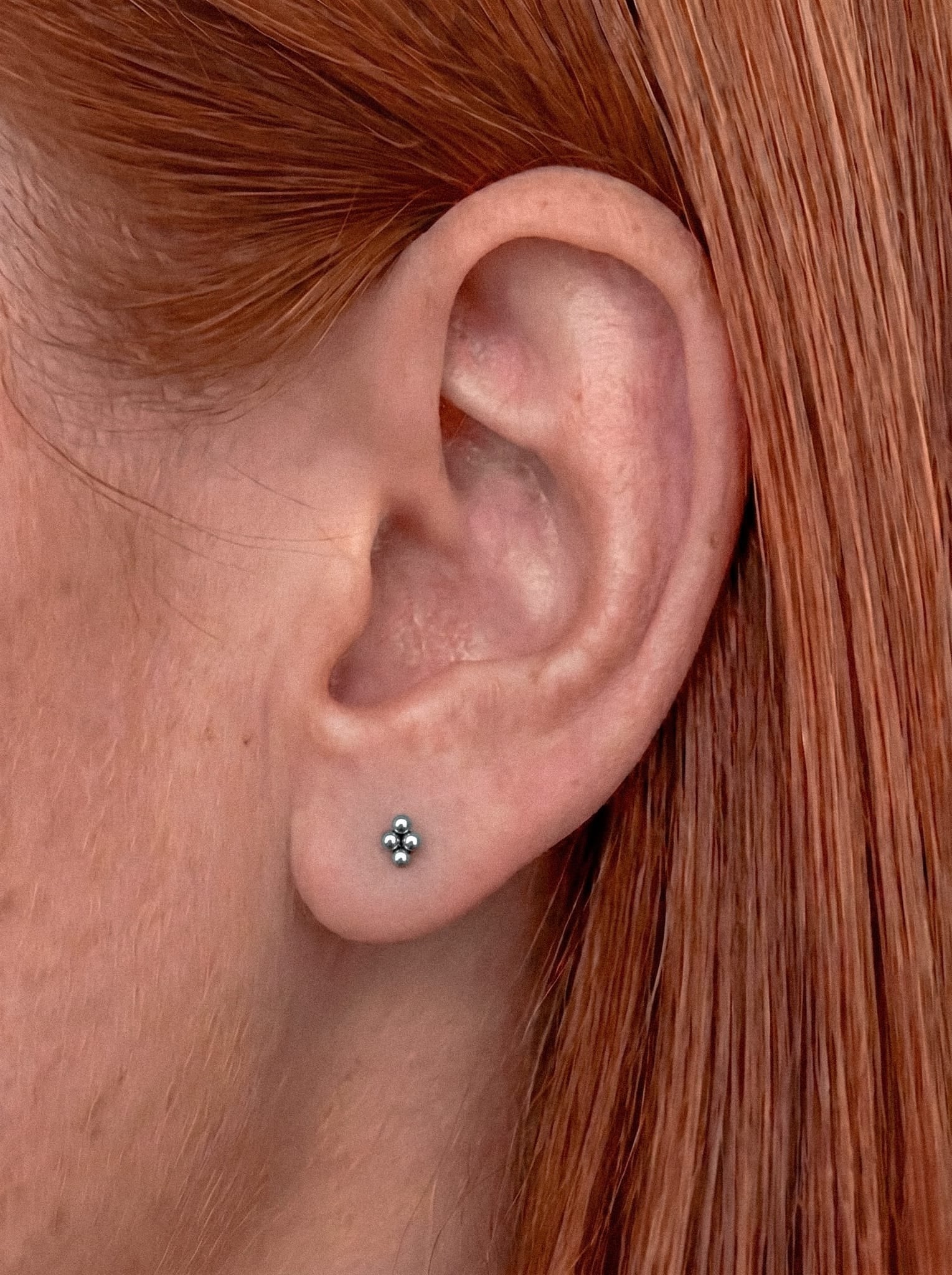Primer plano de la oreja de una persona adornada con el pendiente de botón «Top Nido» de NOBLE ART BEAUTY SOCIEDAD LIMITADA, con tres delicados puntos de plata que aportan un toque elegante sobre un fondo de pelo liso y rojo.