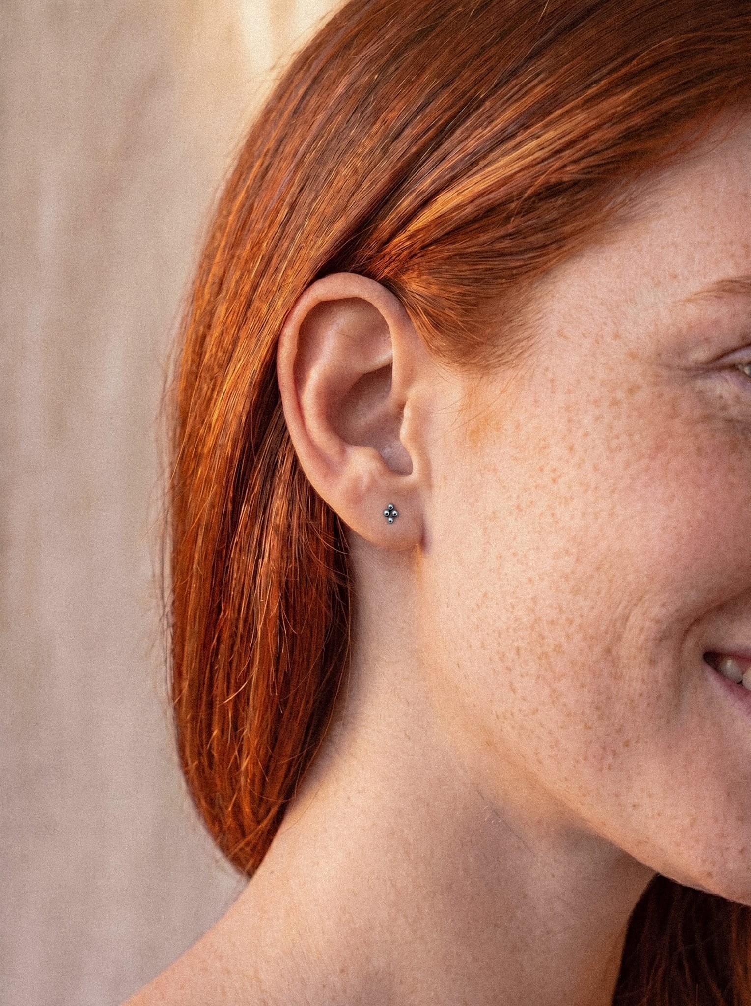 Primer plano de una persona con el pelo largo y pelirrojo y pecas que lleva el «Top Nido», un pequeño pendiente de botón con forma de flor de NOBLE ART BEAUTY SOCIEDAD LIMITADA. La persona sonríe y solo se le ve parte del rostro, lo que capta a la perfección un aspecto delicado, ideal para quienes aprecian los pendientes modernos.