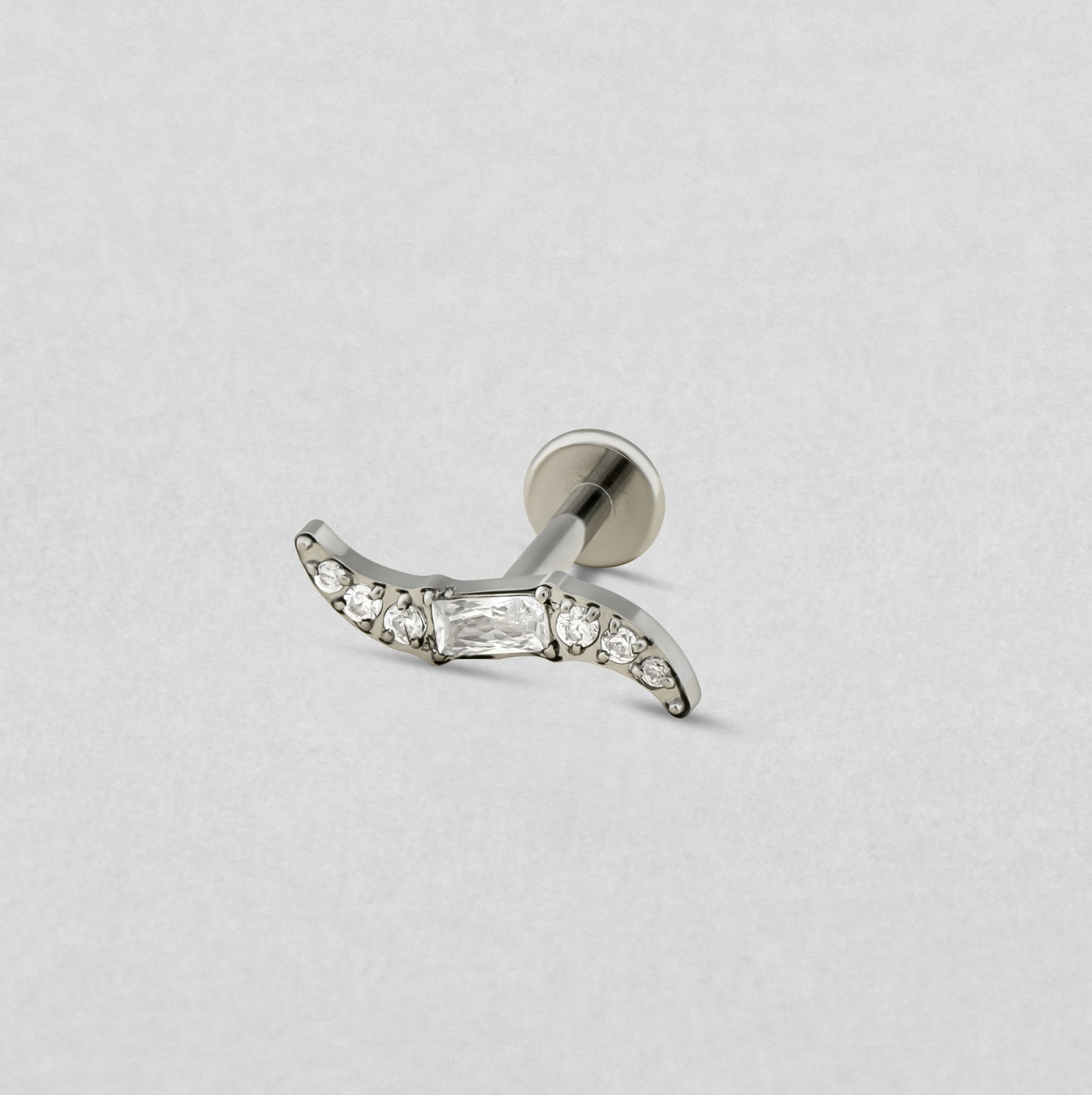 El modelo ONDA SILVER de Titanio-Noble Art es un pendiente de botón de plata con un diseño en forma de media luna, con una pieza central rectangular y pequeñas piedras redondas a lo largo de la curva, que se muestra sobre un fondo liso de color gris claro.