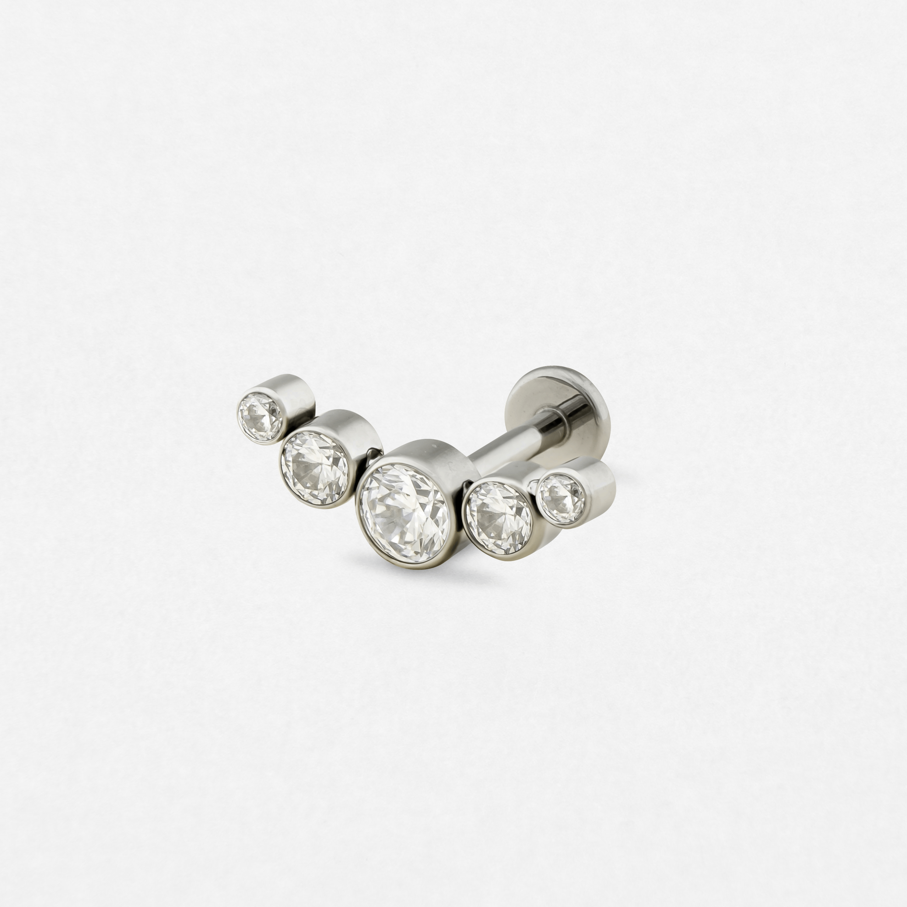 El «Titanio- Noble Art Ornamental 3 cz» es un piercing tipo barra de plata con tres piedras preciosas redondas y transparentes de distintos tamaños dispuestas en forma de curva, que se muestra sobre un fondo blanco.