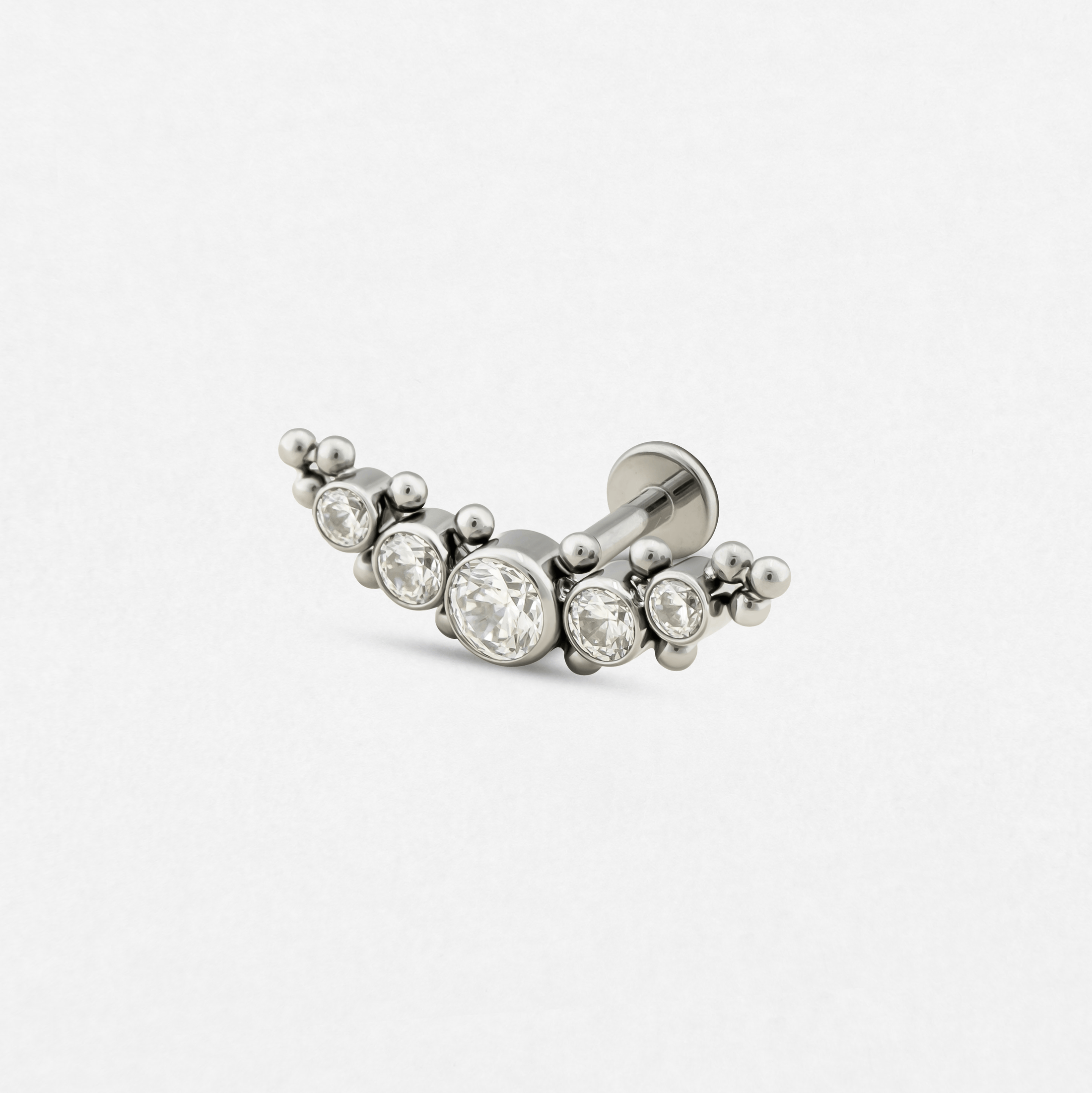 El modelo «Ornamental 5 cz» de la colección Titanio-Noble Art es un pendiente de botón curvo de plata con cinco piedras transparentes de distintos tamaños y pequeños detalles en forma de cuentas, que destacan sobre un fondo blanco liso: un accesorio elegante inspirado en el arte.
