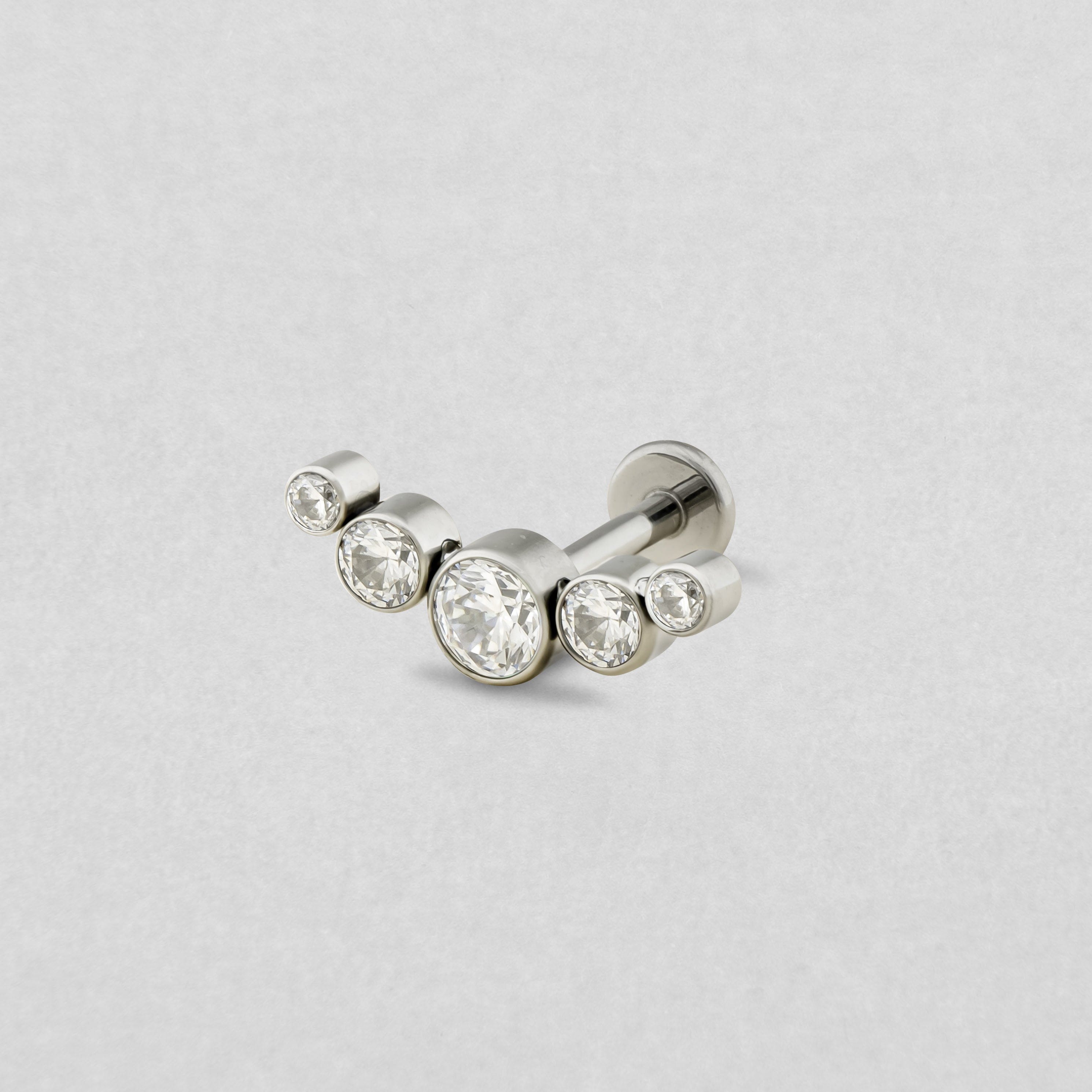 The Titanio–Noble Art "Cluster" model is a silver earring with five clear round cubic zirconia stones arranged in an elegant curve — the largest in the center and smaller ones on either side — displayed against a light gray background.