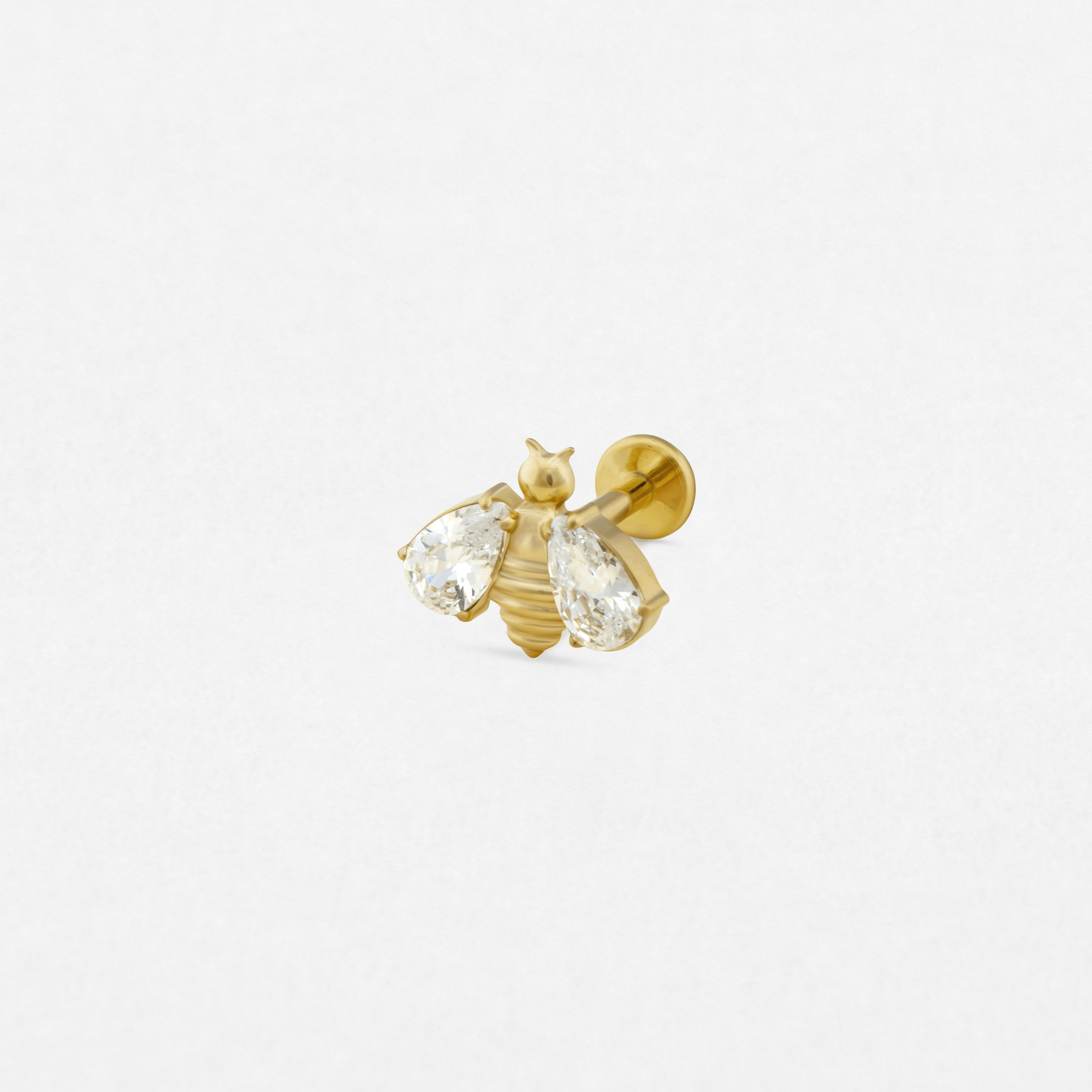 El pendiente de oro «Sol» de Titanio - Noble Art presenta un diseño en forma de abeja con dos piedras preciosas transparentes en forma de lágrima a modo de alas y un cuerpo de oro con textura, lo que aporta elegancia a cualquier piercing. Se muestra sobre un fondo blanco liso.