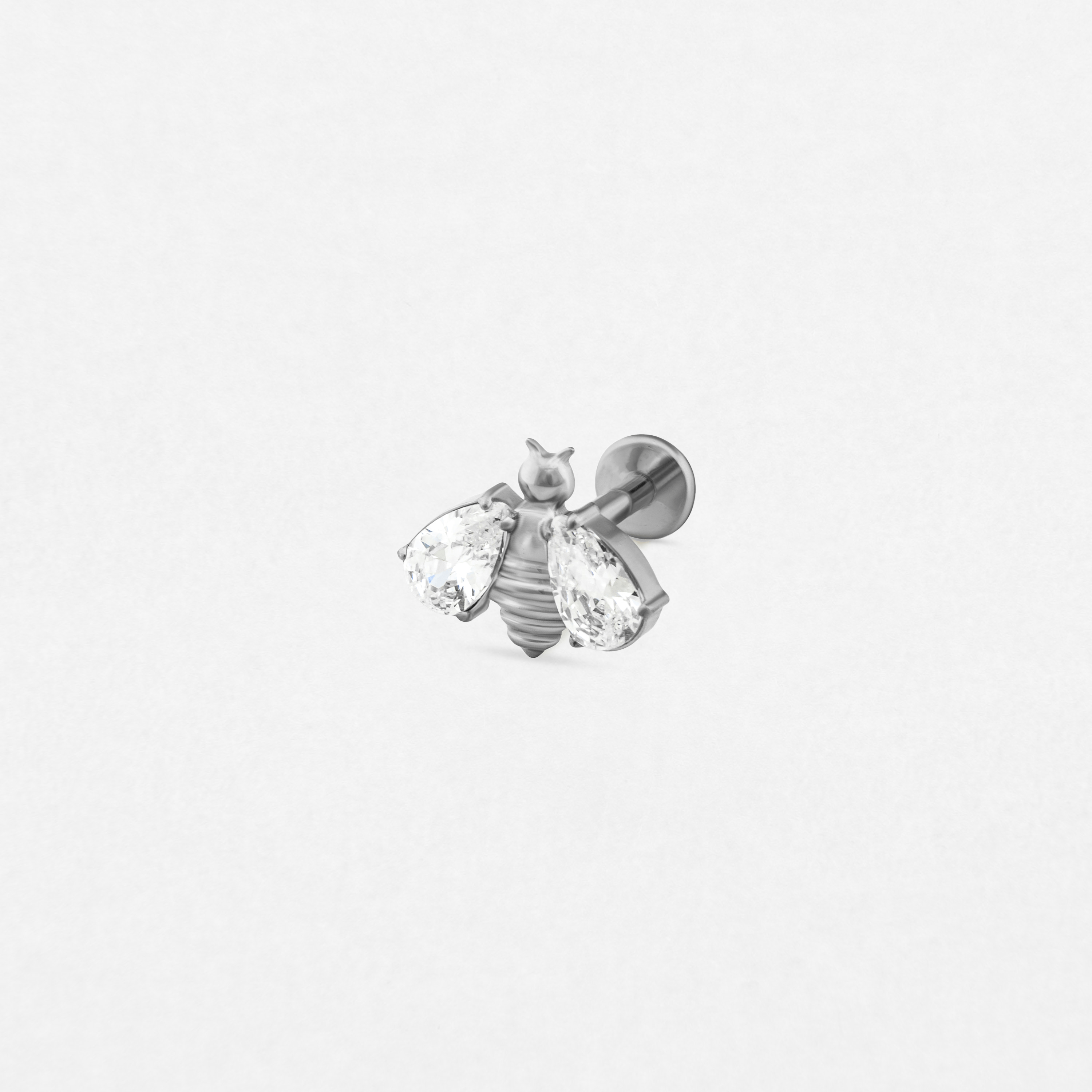 El pendiente «Sol» de Titanio-Noble Art presenta un diseño de abeja en plata con dos gemas transparentes en forma de lágrima a modo de alas, sobre un fondo blanco, ideal para los amantes de la joyería refinada y artística.