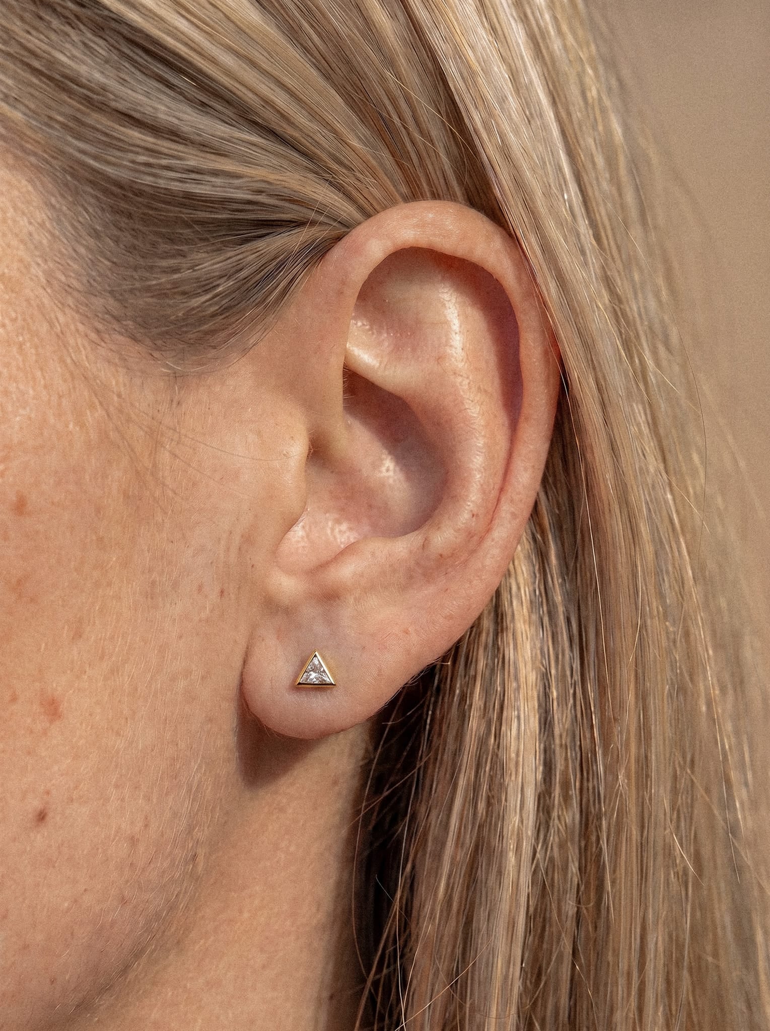 Primer plano de la oreja de una persona que lleva el pendiente de botón «Titin» de Titanio-Noble Art: una pequeña pieza de oro con forma de triángulo. La persona tiene la piel clara y el pelo liso y rubio.