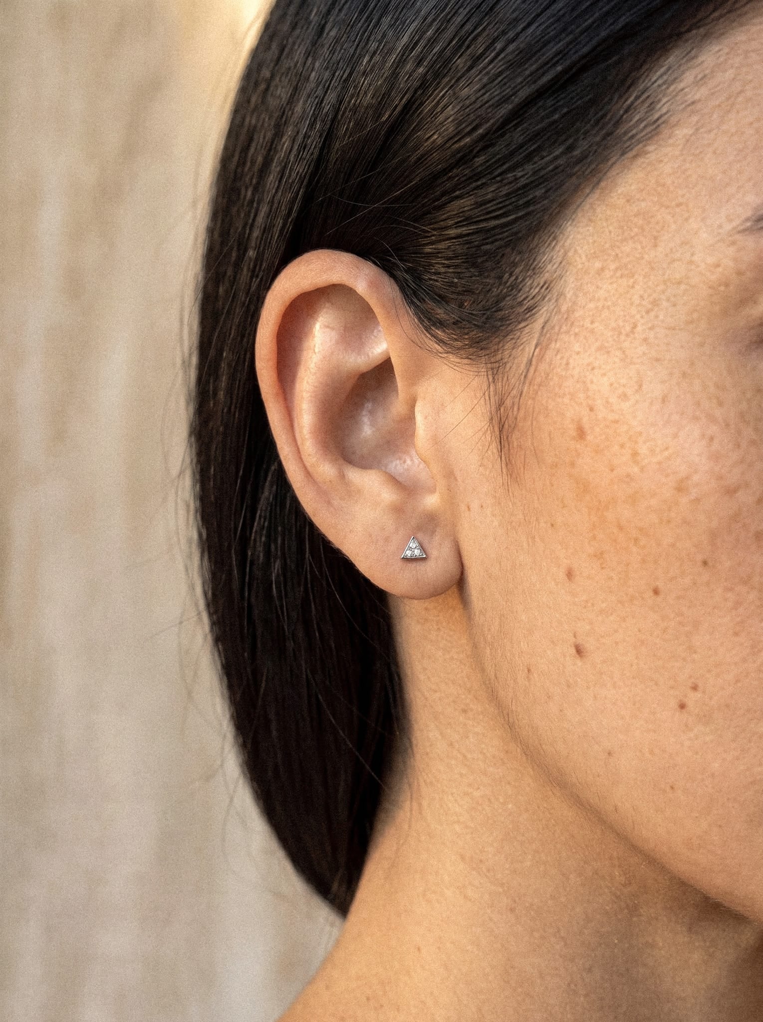 Primer plano de la oreja de una mujer con el pelo liso y oscuro, que luce el pendiente triangular de plata «Titin» de Titanio-Noble Art. Su piel clara, salpicada de algunas pecas y lunares, destaca sobre un fondo suavemente difuminado de tonos neutros.