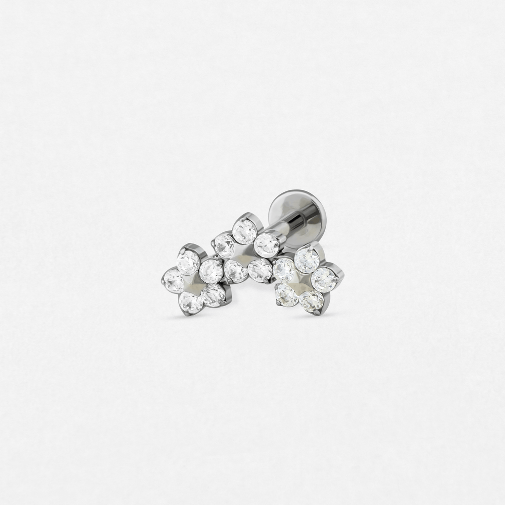 The 'Titanio-Noble Art Triple Flower' is a silver stud earring with three clusters of small round clear stones in a floral design, ideal for adding subtle elegance to any piercing, as shown against a plain white background.
