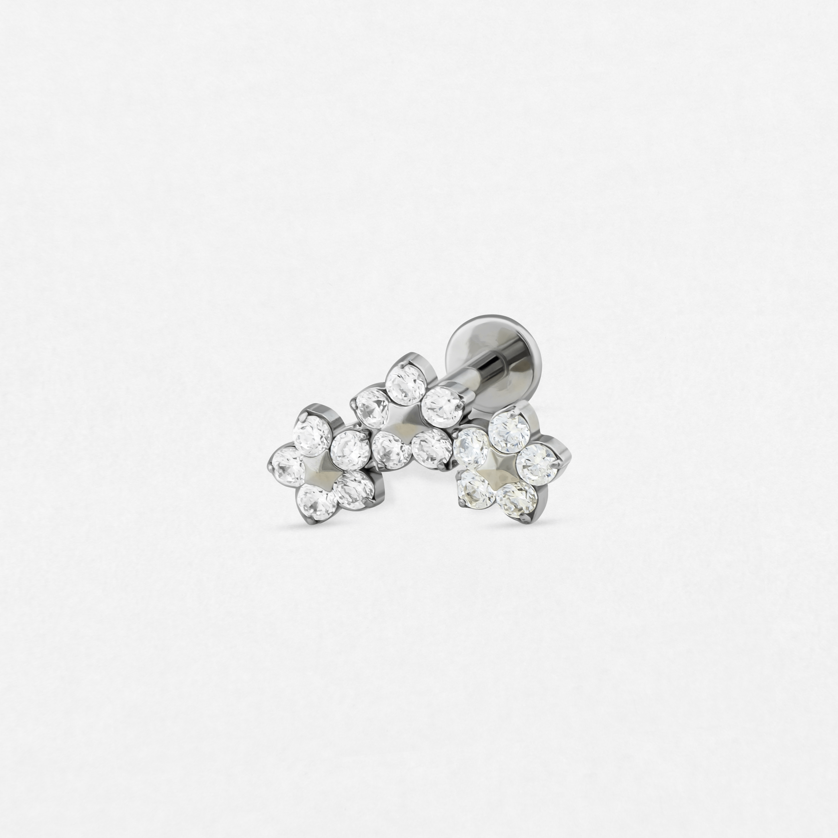 El modelo «Titanio- Noble Art Triple Flower» es un pendiente de botón de plata con tres racimos de pequeñas piedras redondas transparentes dispuestas en un diseño floral, ideal para aportar una elegancia sutil a cualquier piercing, tal y como se muestra sobre un fondo blanco liso.