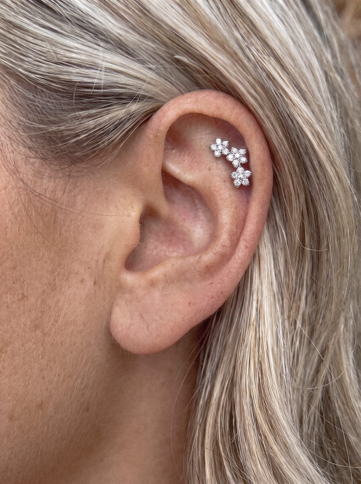 Primer plano de la oreja de una persona adornada con los pendientes de diamantes «Triple flower» de Titanio-Noble Art, que cuentan con tres pequeños pendientes en forma de flor a lo largo del cartílago del hélix. El pelo rubio claro de la persona realza este elegante y floral look de piercing.