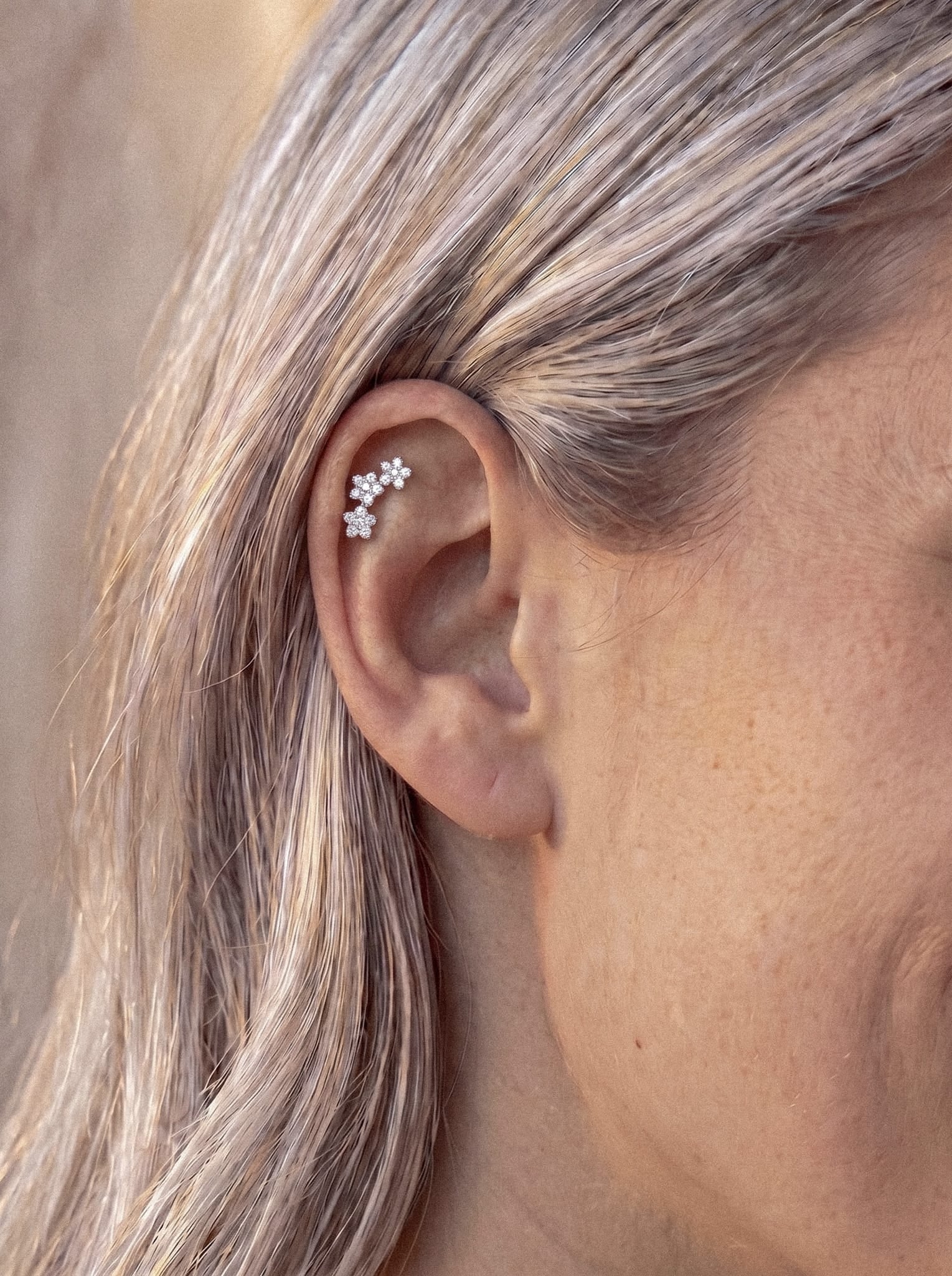 Primer plano de la oreja de una persona con el pelo rubio claro, que luce el pendiente tipo «ear cuff» de plata «Triple flower» de Titanio-Noble Art, delicadamente adornado con tres pequeños motivos florales que le aportan un toque elegante, perfecto para resaltar el hélix o el trago y realzar cualquier look de inspiración floral.