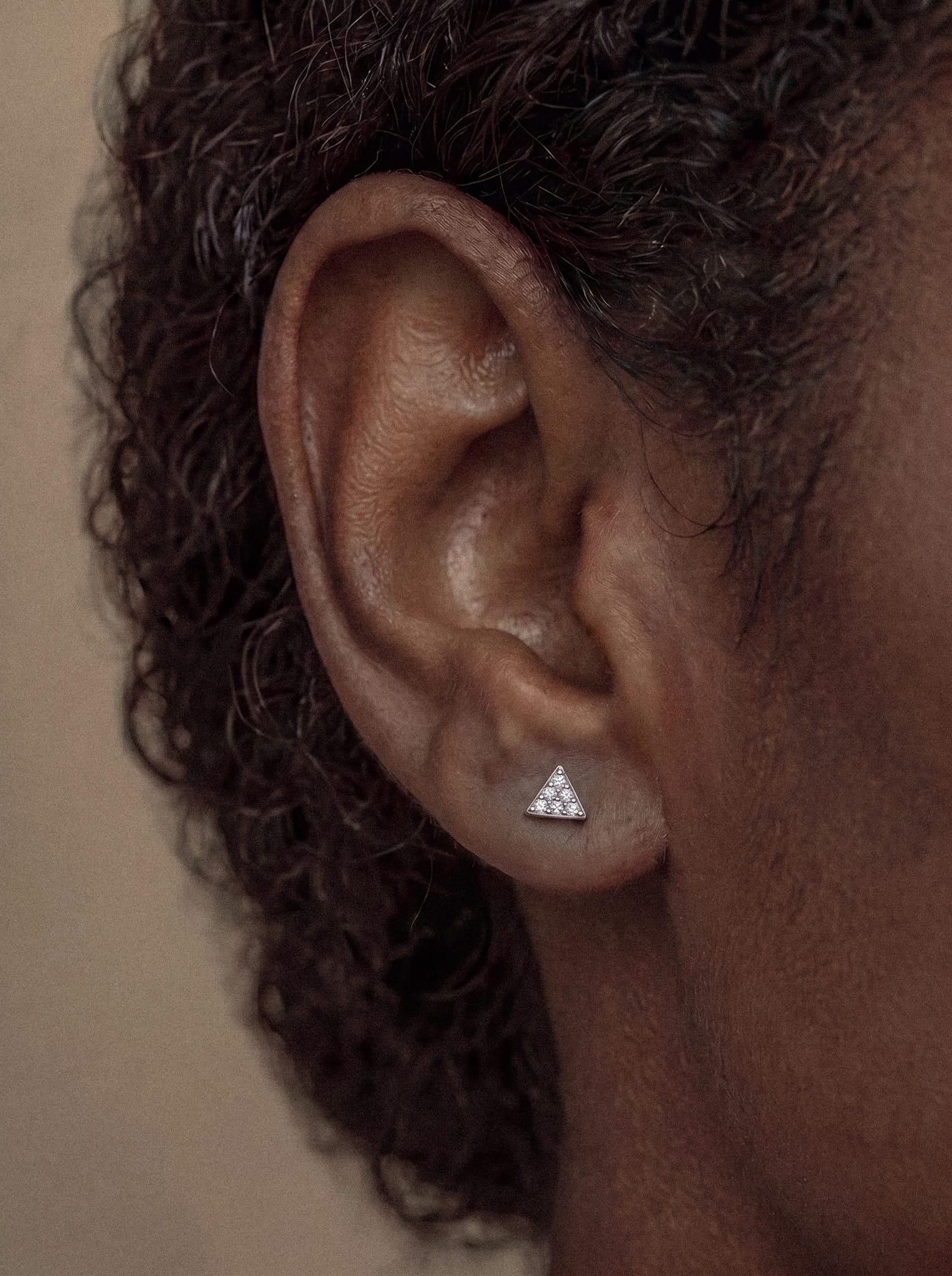 Primer plano de la oreja de una persona que lleva el pendiente de botón «Trianglue CZ» de Titanio-Noble Art, un pequeño pendiente de plata con forma triangular y un cristal brillante. La piel de la persona es de tono moreno medio y su pelo rizado se vislumbra suavemente al fondo. Esta pieza encarna la joyería minimalista con sus líneas limpias y su diseño moderno.