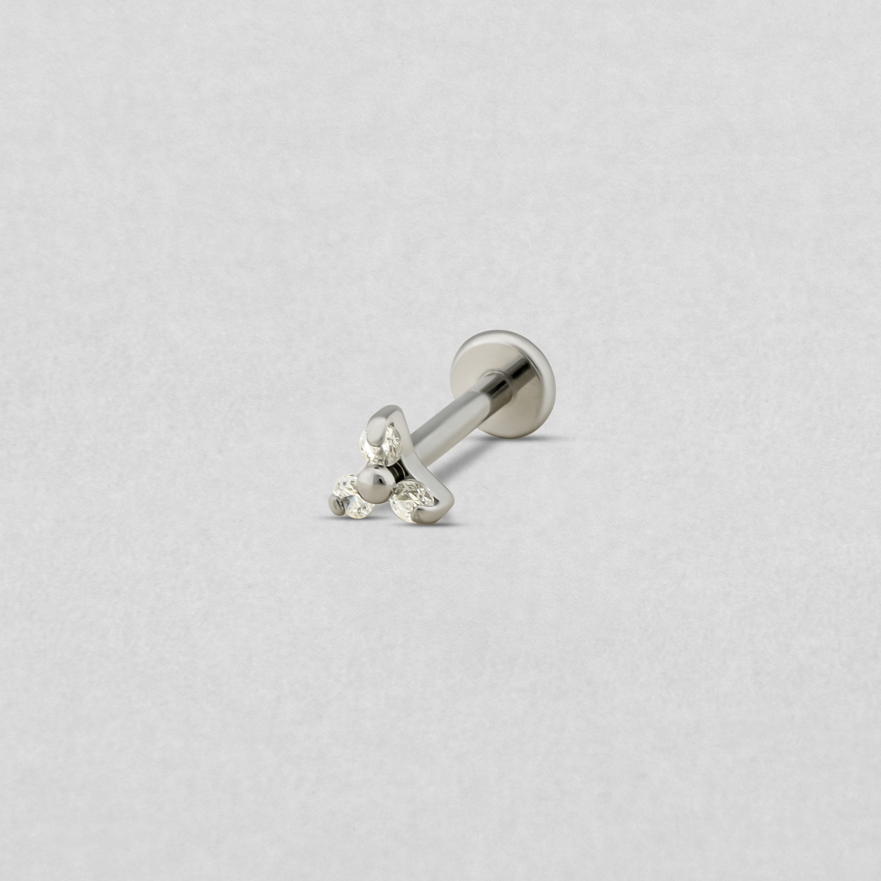 El «Trinity cz» de Titanio-Noble Art es un pendiente labret de plata con la parte trasera plana y un conjunto de tres piedras transparentes en la parte delantera, que se muestra sobre un fondo gris claro; ideal para los seguidores de Noble Art tras un tatuaje o una eliminación con láser.