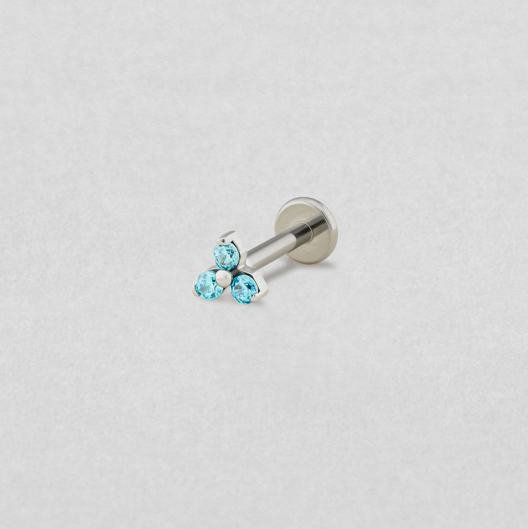El pendiente de labret «Trinity cz» de Titanio-Noble Art cuenta con tres pequeñas piedras preciosas redondas de color azul claro dispuestas en forma de triángulo, que se muestran sobre un fondo blanco liso.