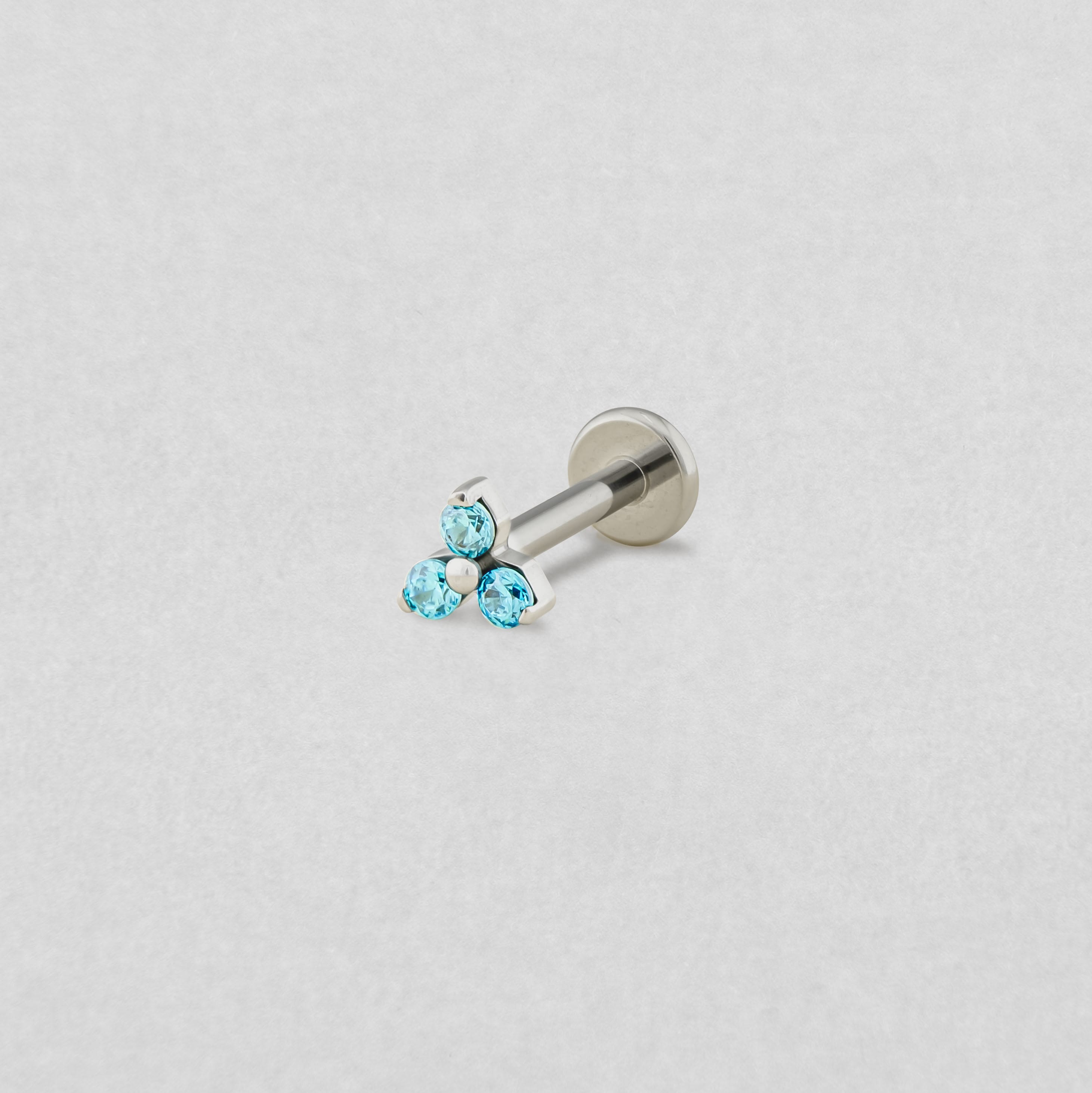 El pendiente de labret «Trinity cz» de Titanio-Noble Art cuenta con tres pequeñas piedras preciosas redondas de color azul claro dispuestas en forma de triángulo, que se muestran sobre un fondo blanco liso.