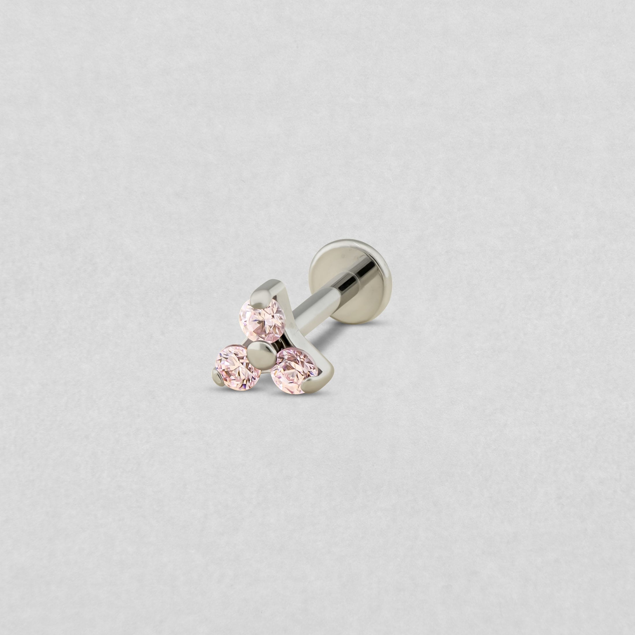 El «Trinity cz» de Titanio-Noble Art es un pendiente tipo labret de plata con tres piedras preciosas de color rosa claro dispuestas en forma de triángulo, ideal para cualquier tipo de piercing y presentado sobre un fondo gris claro.