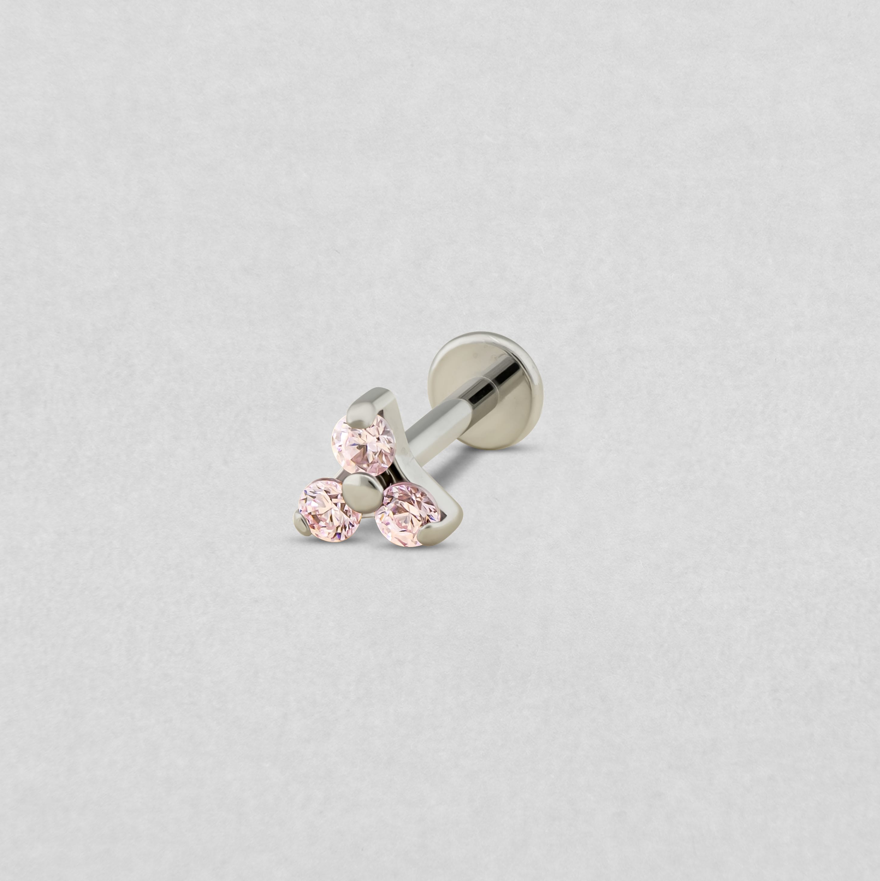El «Trinity cz» de Titanio-Noble Art es un pendiente tipo labret de plata con tres piedras preciosas de color rosa claro dispuestas en forma de triángulo, ideal para cualquier tipo de piercing y presentado sobre un fondo gris claro.