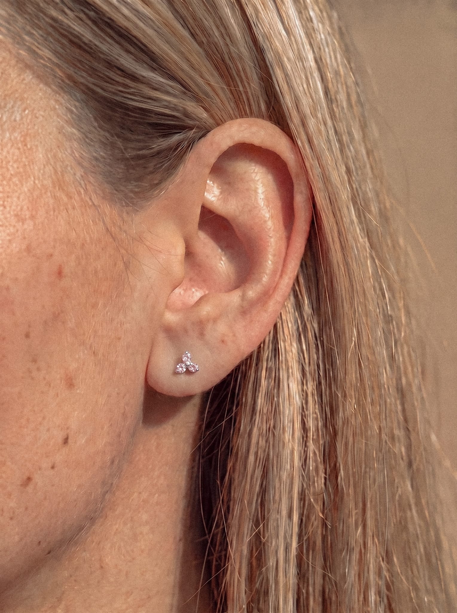 Nahaufnahme des Ohrs einer Person mit glattem, hellbraunem Haar, die den Ohrstecker „Trinity cz“ von Titanio-Noble Art trägt – ein kleines, blumenförmiges Design, verziert mit funkelnden Kristallen. Die Haut ist hell und der Hintergrund ist sanft unscharf.