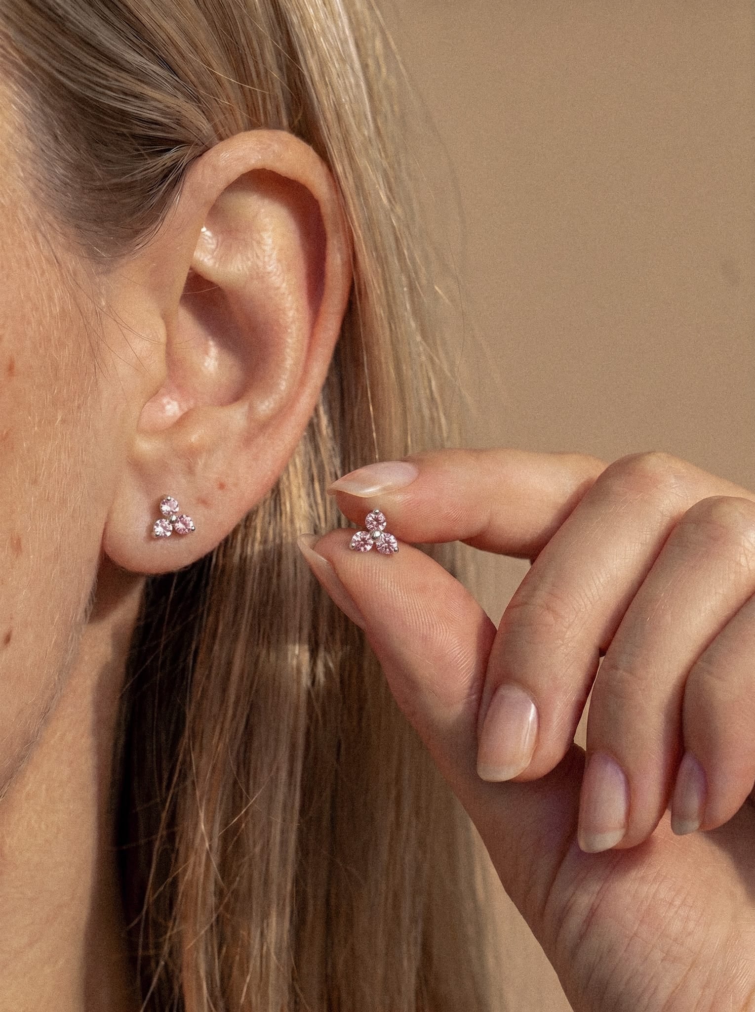 Eine Nahaufnahme des Ohrs einer Person zeigt den Ohrring „Trinity cz“ von Titanio-Noble Art, der sich durch ein zierliches, blumenförmiges Design in Rosa auszeichnet, das mit funkelnden Kristallen besetzt ist. Die Person hält einen passenden Ohrring behutsam an ihr Ohr und unterstreicht so den dezenten Glanz dieses eleganten Accessoires. Der Hintergrund ist neutral gehalten, und die Haare sind ordentlich hinter dem Ohr gesteckt.