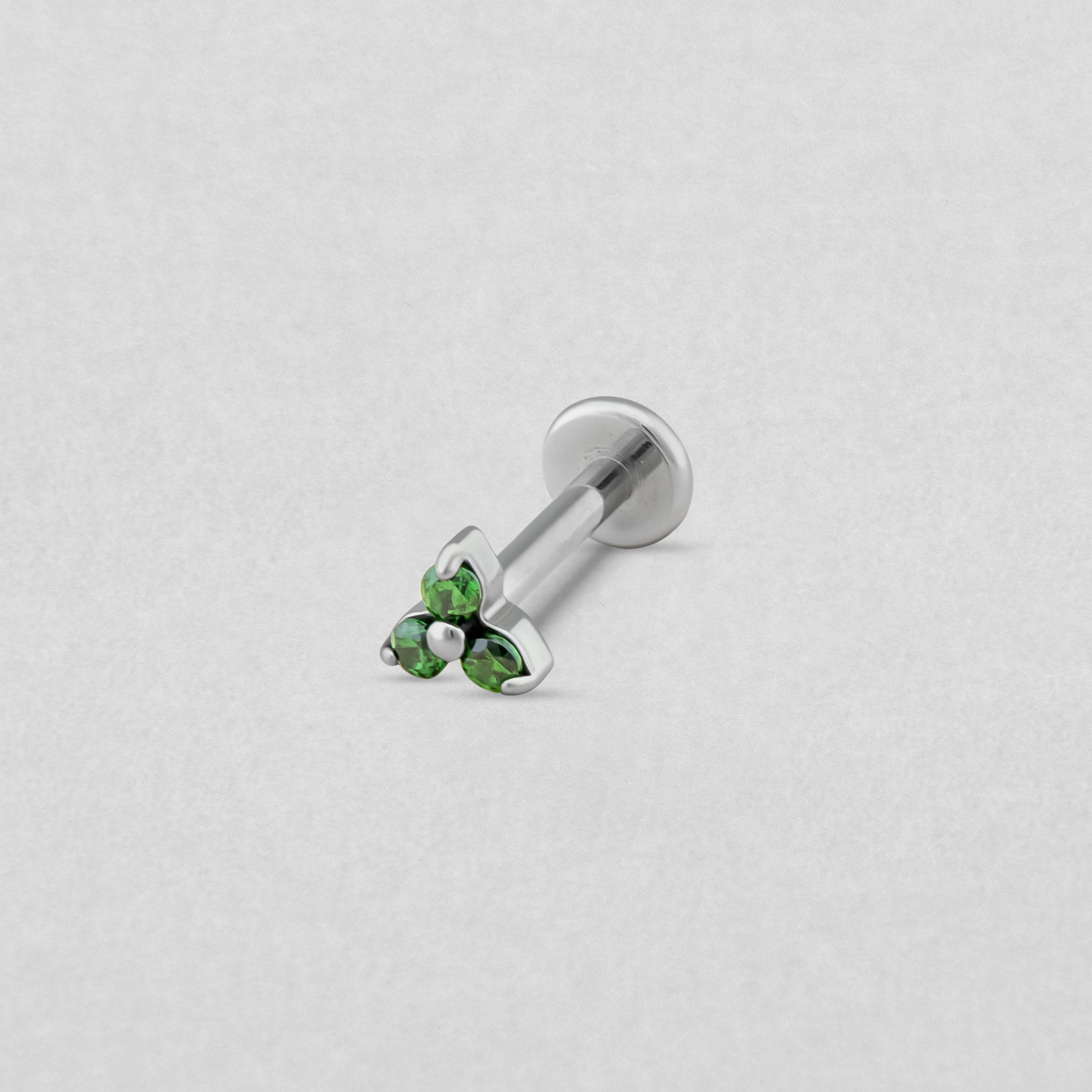 El «Trinity cz» de Titanio-Noble Art es un pendiente tipo labret de plata para piercings, que presenta un conjunto triangular de piedras preciosas verdes en un extremo, elegantemente resaltadas sobre un fondo texturizado de color gris claro.